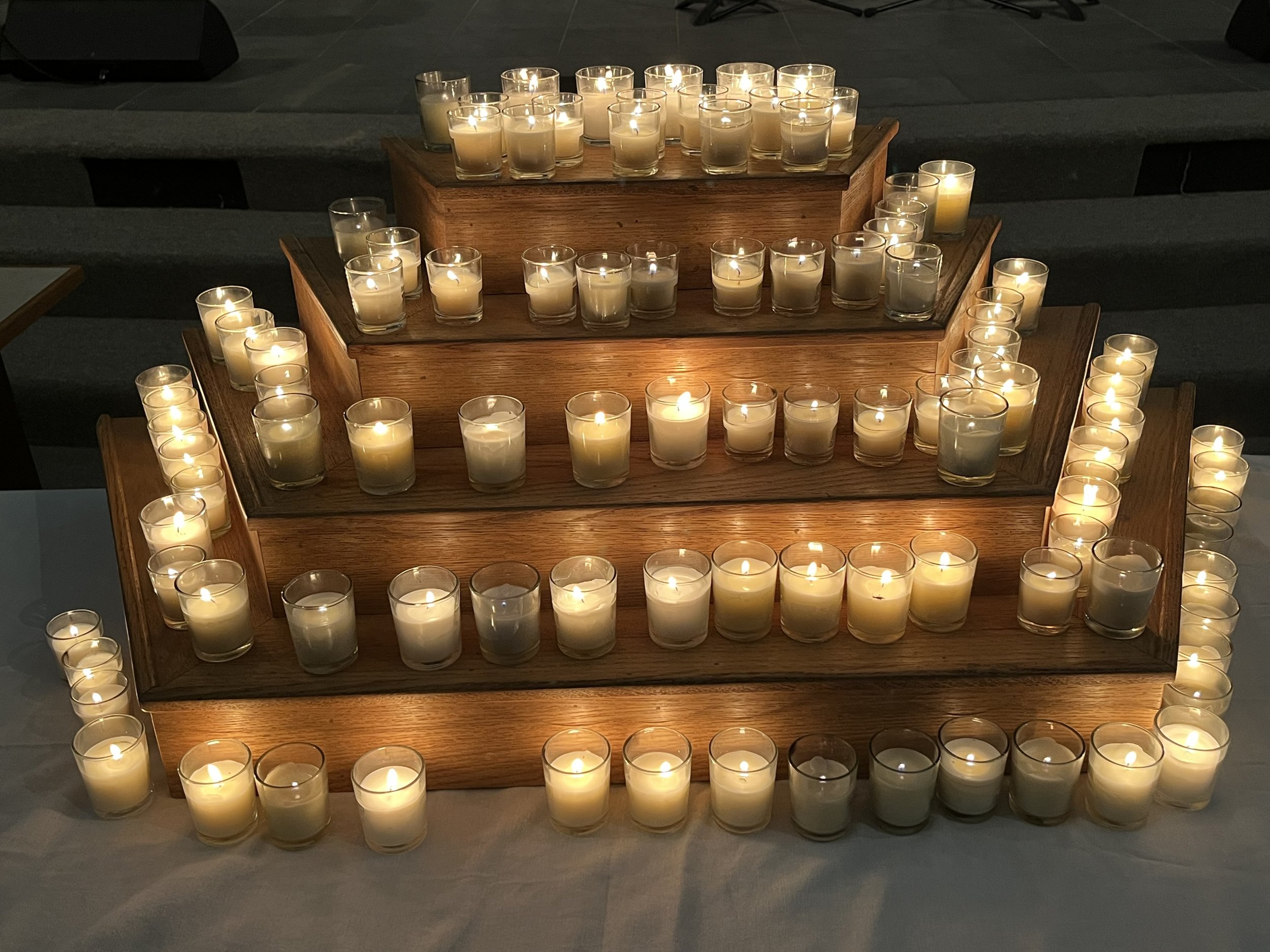 A multi-tiered wooden display with numerous lit candles in glass holders.