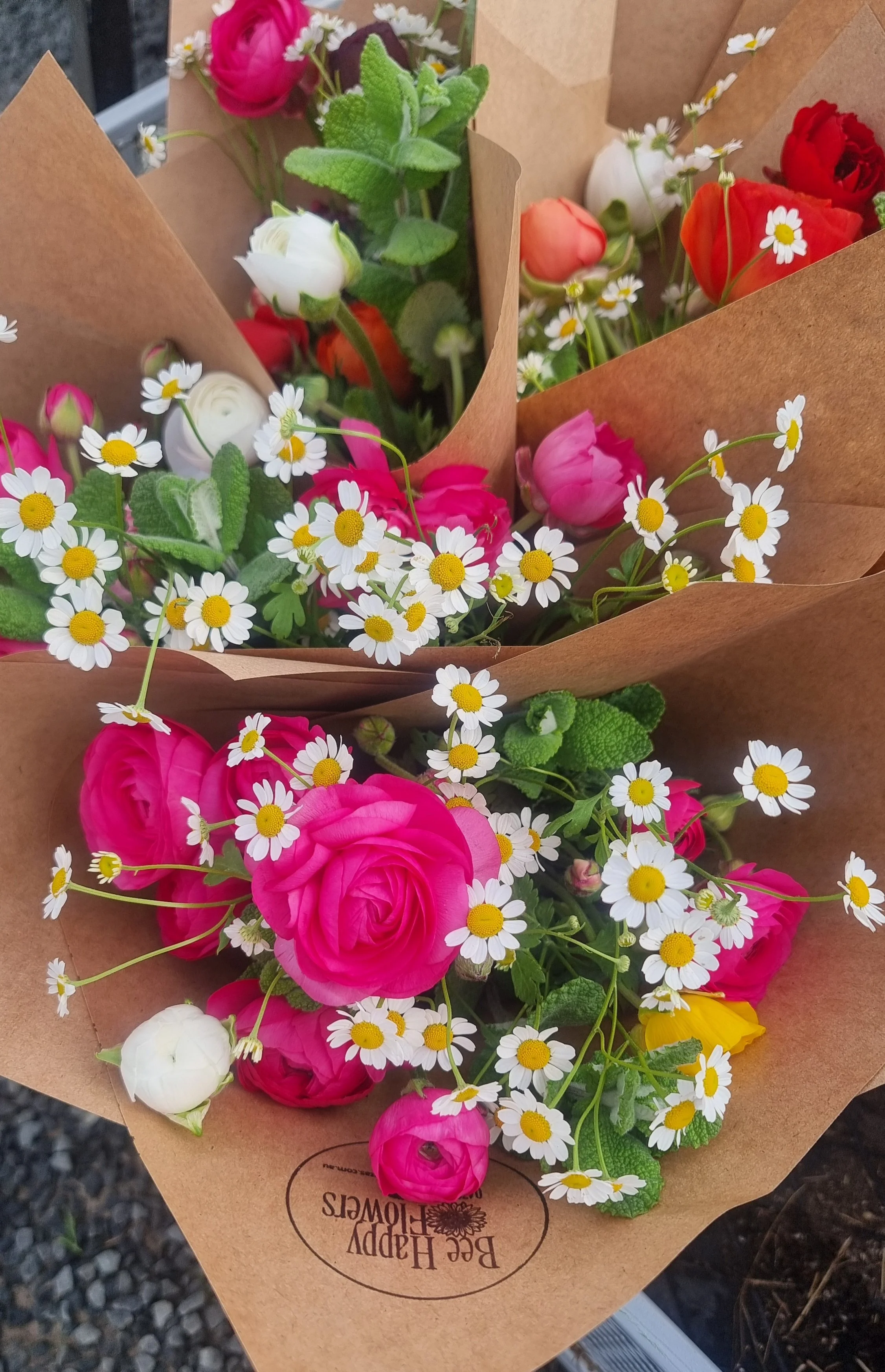 Bouquet of pink roses, white ranunculus, and daisies wrapped in brown paper with "Bee Happy Flowers" logo.