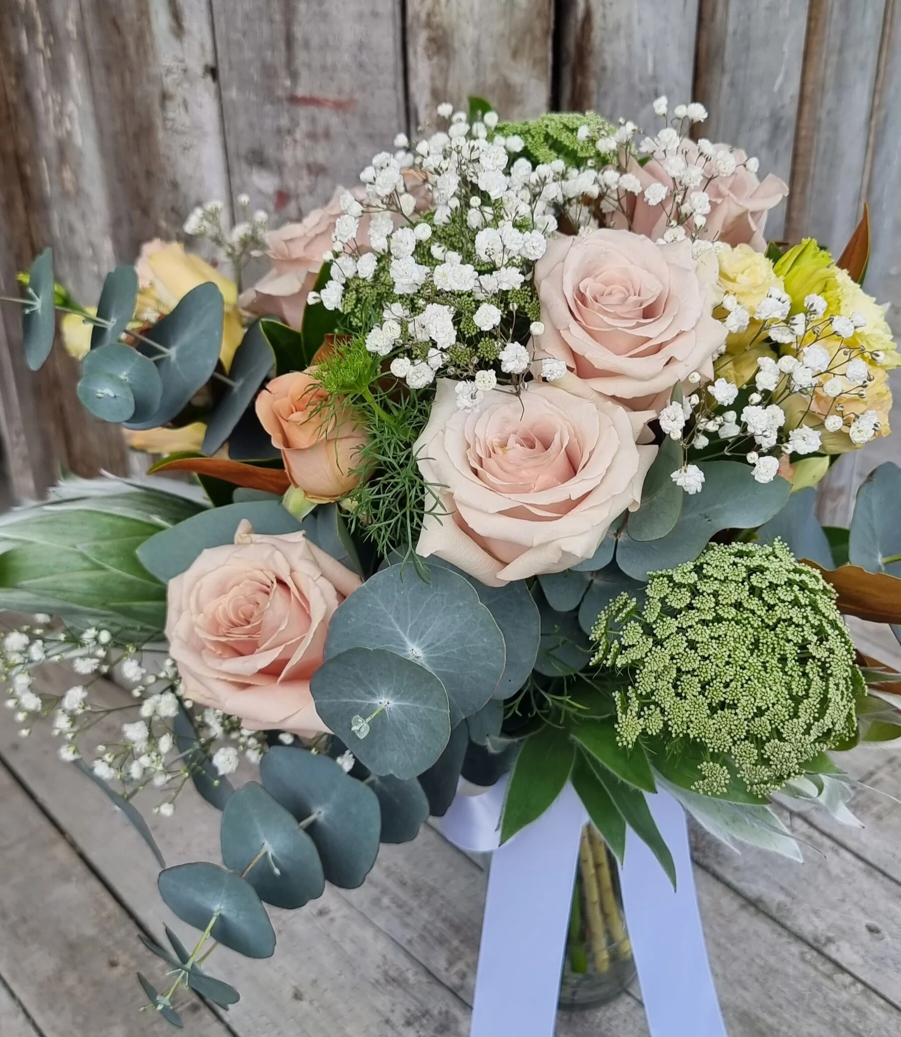 A bouquet of pink roses, baby's breath, eucalyptus, green filler flowers, and assorted greenery in a glass vase with a blue ribbon, on a rustic wooden surface.