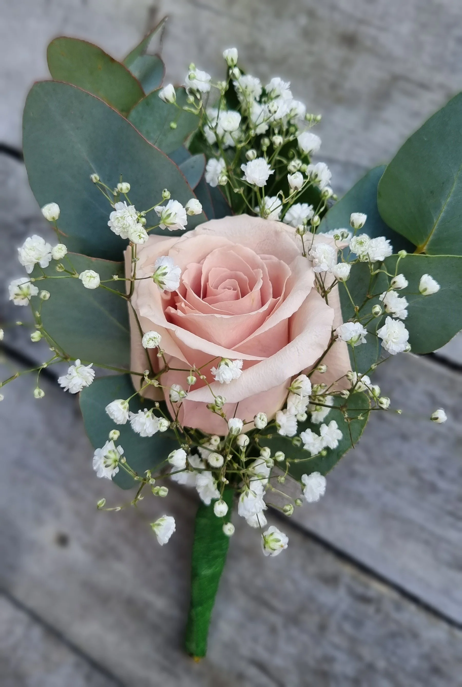 A boutonniere with a pale pink rose, surrounded by white baby's breath flowers and green eucalyptus leaves on a wooden surface.