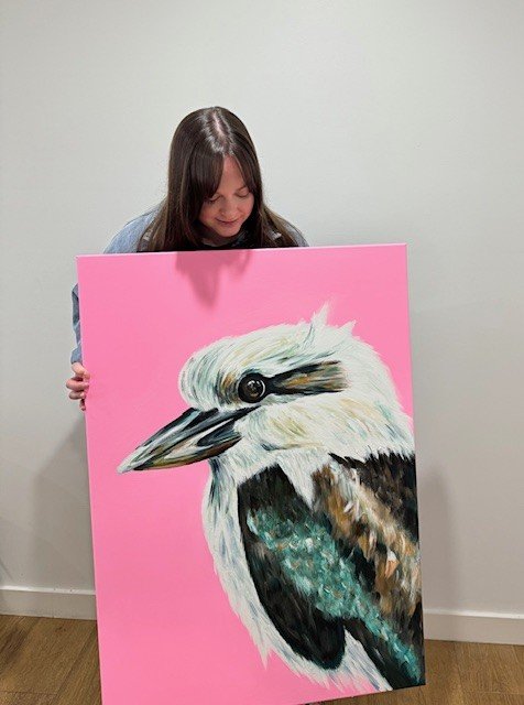 Laura Boatman holding a large painting of a colorful bird with a pink background. A commissioned art piece.