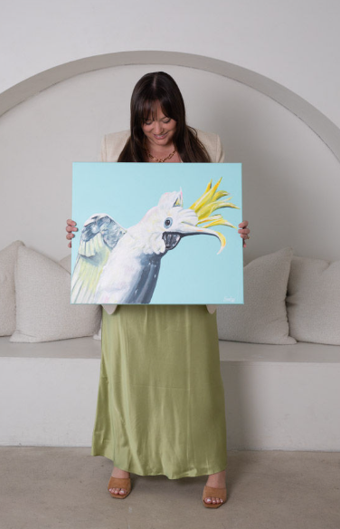 Woman holding a painting of a bird with a beak resembling a banana, in a white room with a curved wall and cushions.