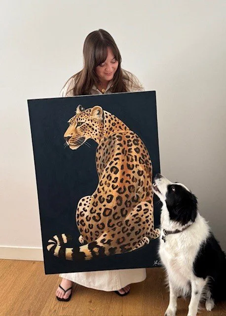 A woman holding a large painting of a leopard sitting, with a black and white dog sitting nearby and looking at it.