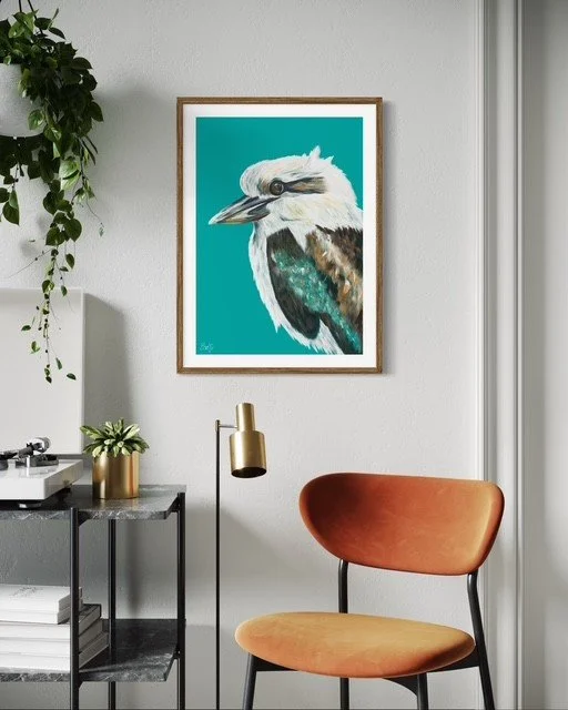 A framed art print of a koala bird is hung on a white wall, with a green potted plant on a small table below it. There is a modern floor lamp with a gold shade and an orange chair with a black metal frame in front of the wall.