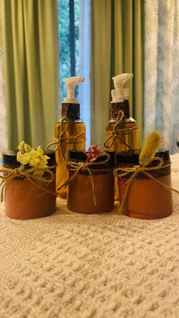 Three brown jars with twine bows and decorative flowers, and two amber spray bottles with white spray nozzles, arranged on a textured beige surface in front of curtains and a window.