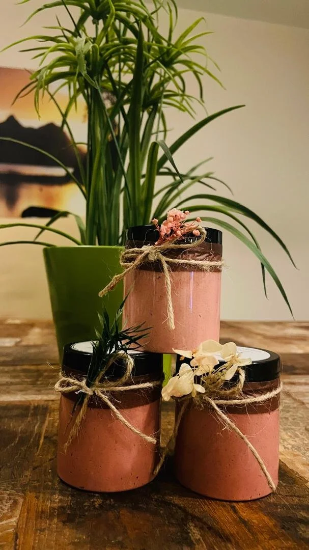 Three pink cylindrical jars with black lids, tied with twine bows, are displayed on a wooden surface, with various dried flower decorations attached. A tall green potted plant with long leaves is in the background.