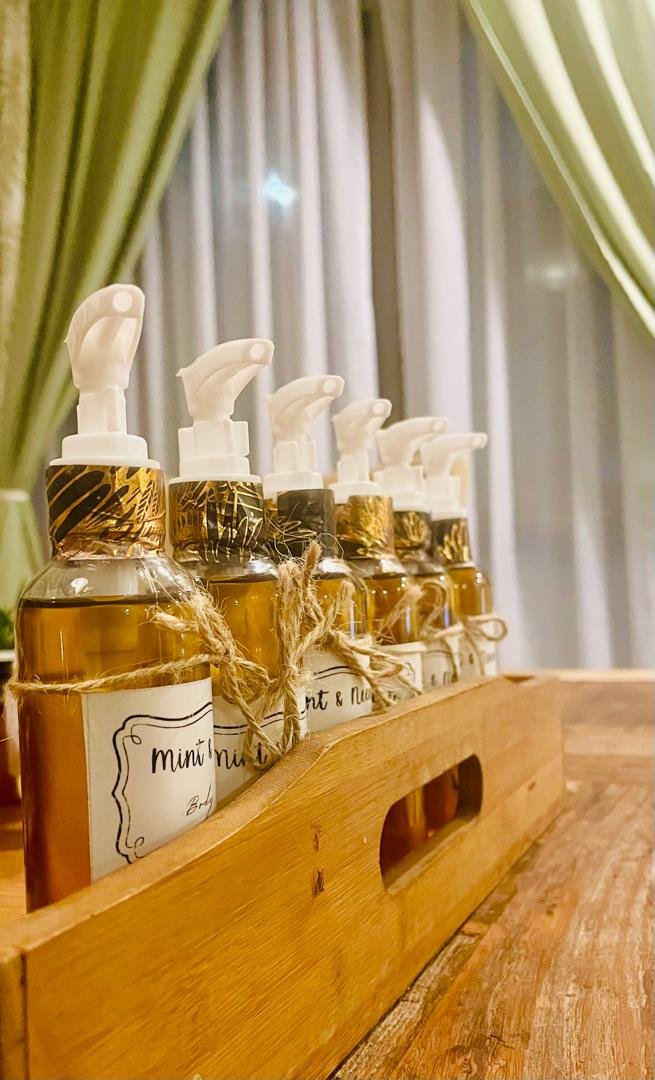 Six amber-colored spray bottles with white nozzles and gold accents, labeled 'Mint & Nut' and tied with twine, are arranged in a wooden caddy on a wooden surface, with green curtains and white sheer curtains in the background.