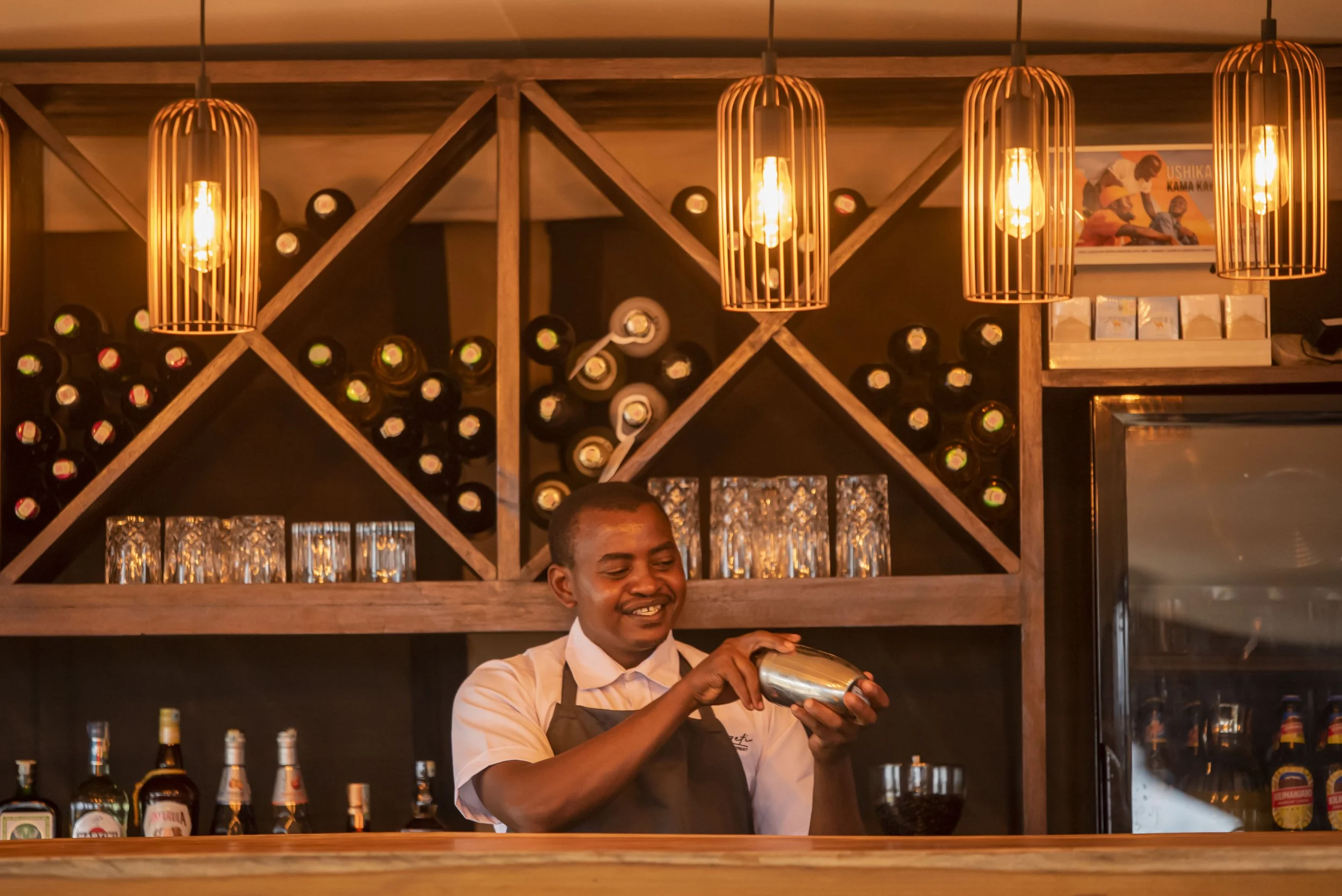 A bartender smiling while preparing a drink in a bar with warm lighting, wine bottles on shelves, glasses, and artwork on the wall.