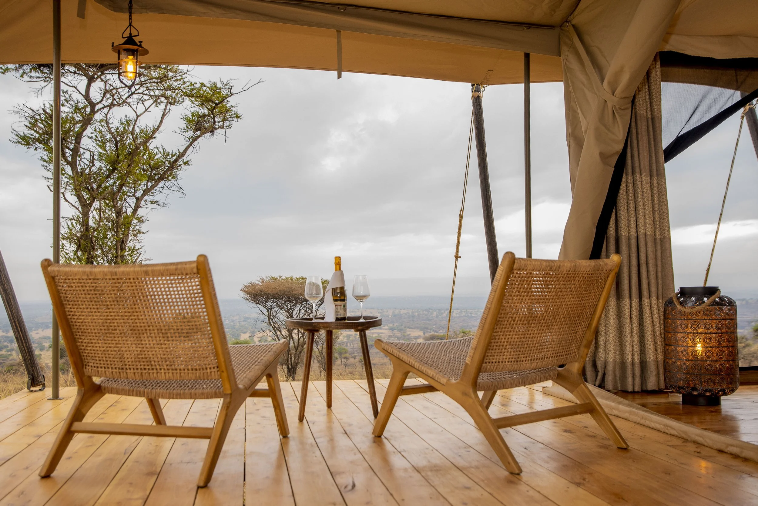 A scenic outdoor dining area with two woven chairs, a small round wooden table set with a bottle of champagne and two glasses, situated on a wooden deck under a canopy. The background features a view of trees and a cloudy sky.