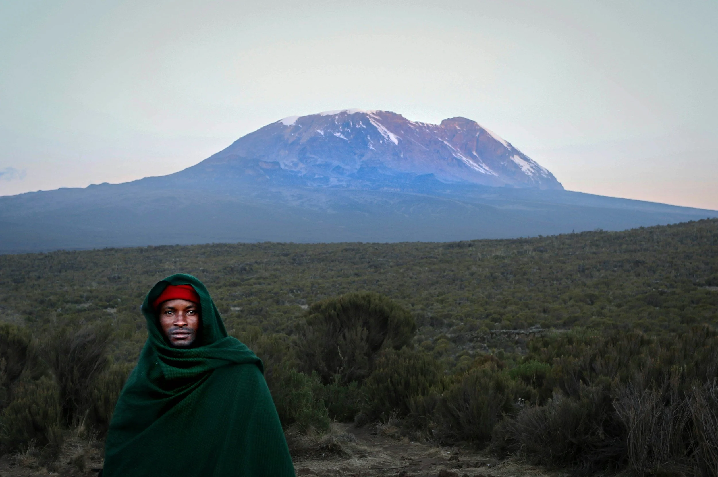 A man dressed in traditional Maasai clothing with a green shuka and red headband, standing in a grassy landscape with Mount Kilimanjaro in the background.
