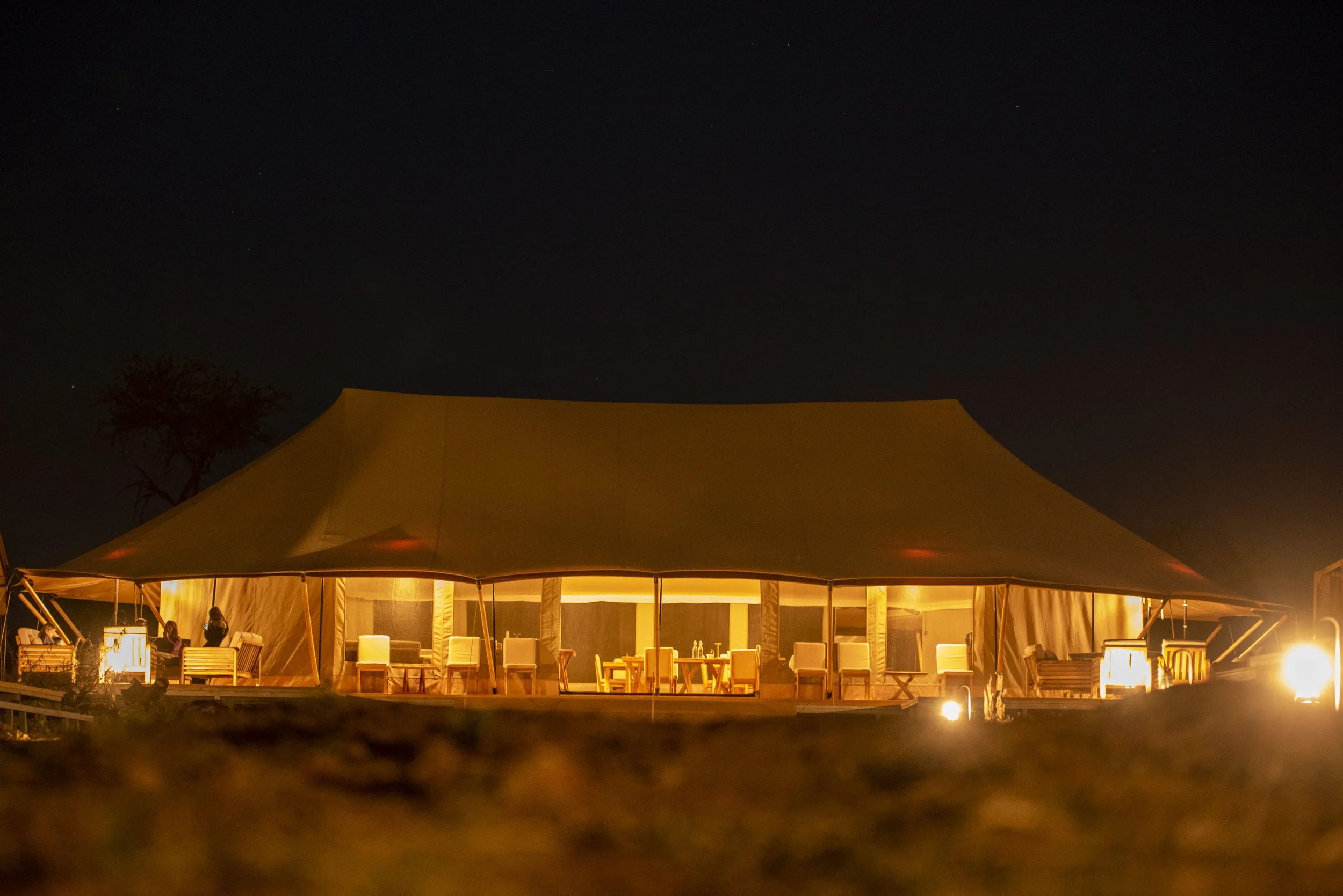 Large tent with warm interior lighting at night, surrounded by chairs and tables, outdoor setting.