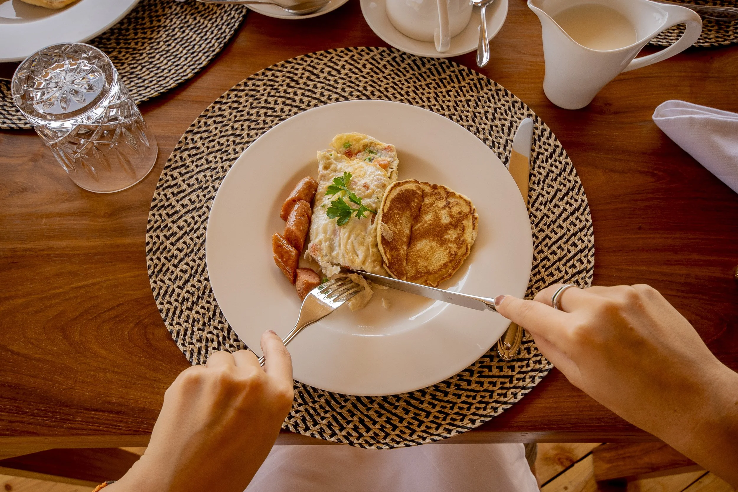 A plate of breakfast food including scrambled eggs, sausage links, and pancakes, with a sprig of parsley, on a woven placemat at a wooden table, with utensils, a glass of water, a gravy boat, and a creamer pitcher nearby.
