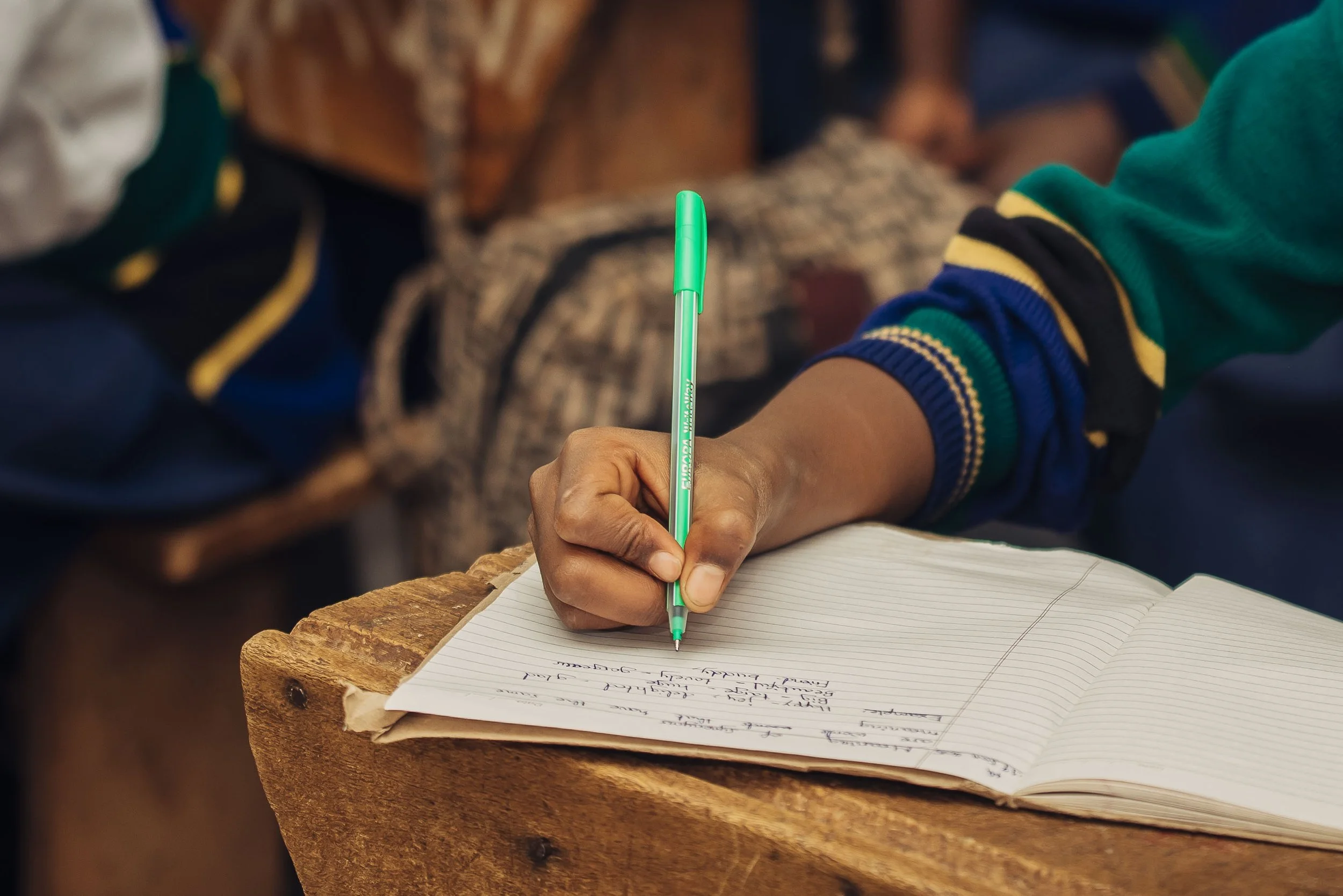 A person writing in a notebook with a green pen on a wooden surface.
