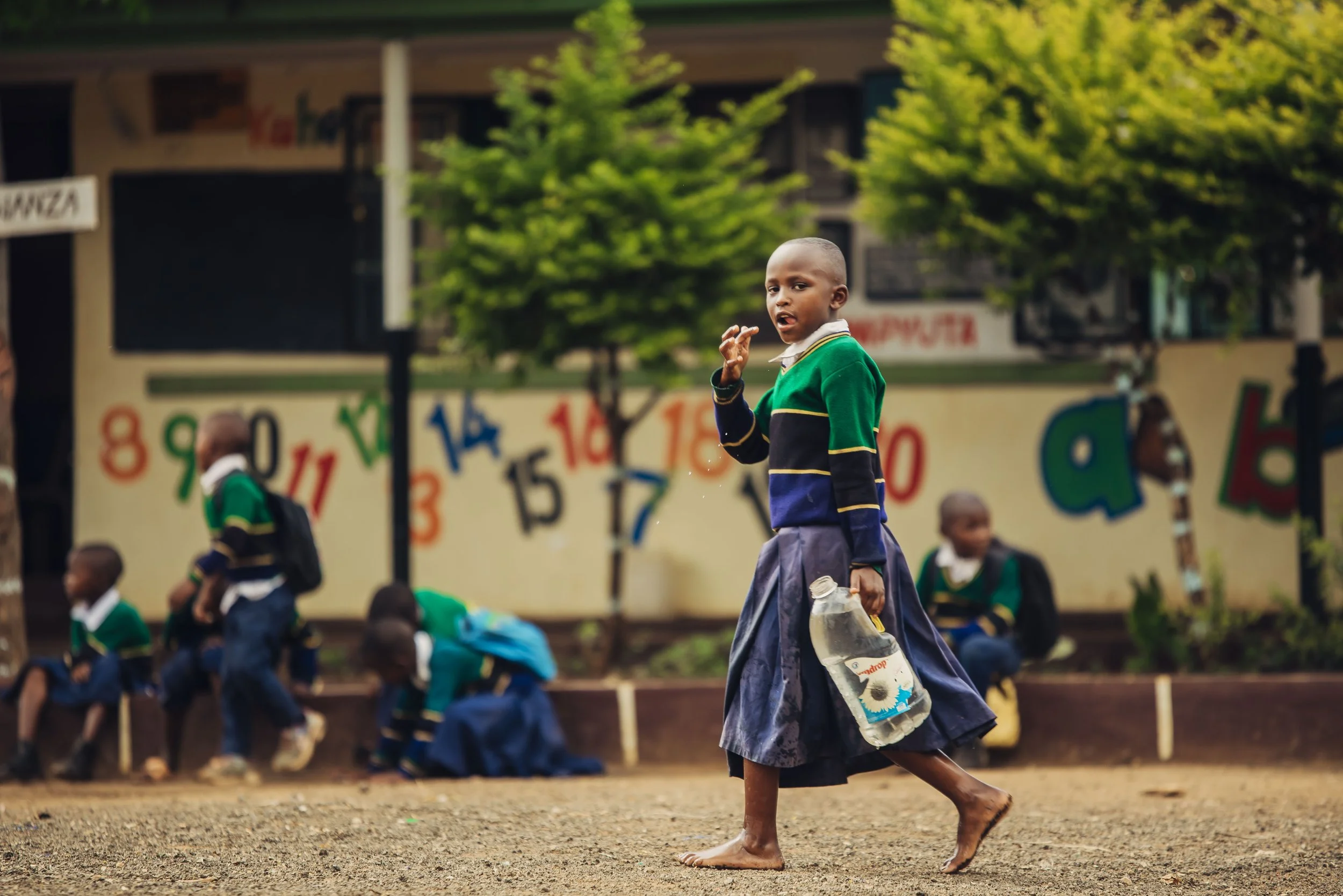 A young boy walking barefoot on a dirt ground holding a plastic bottle, with several children in school uniforms sitting and standing in the background, and colorful numbers painted on a wall behind them.