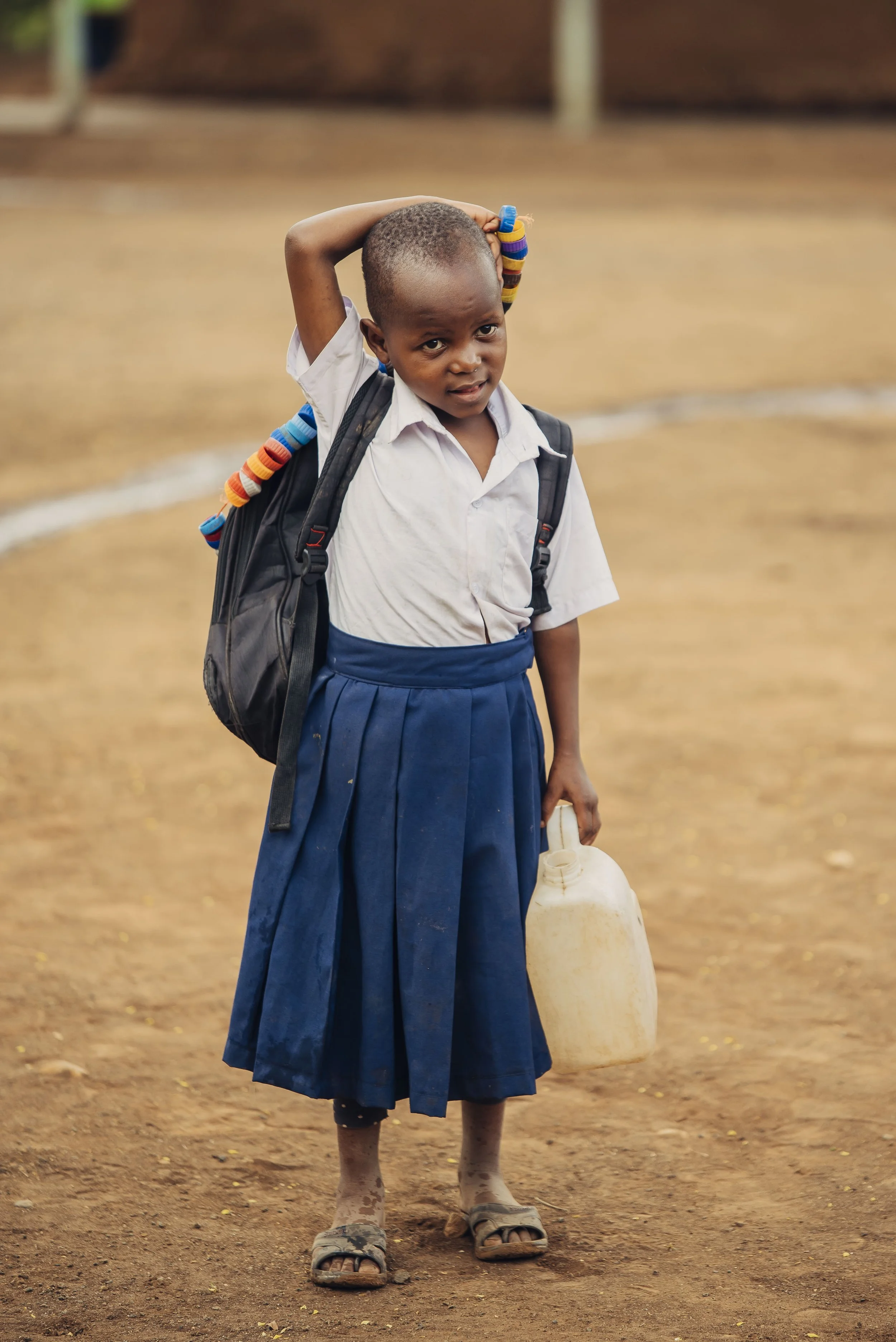 A young girl in school uniform holding a container, with a backpack and a colorful accessory, standing on a dirt schoolyard.