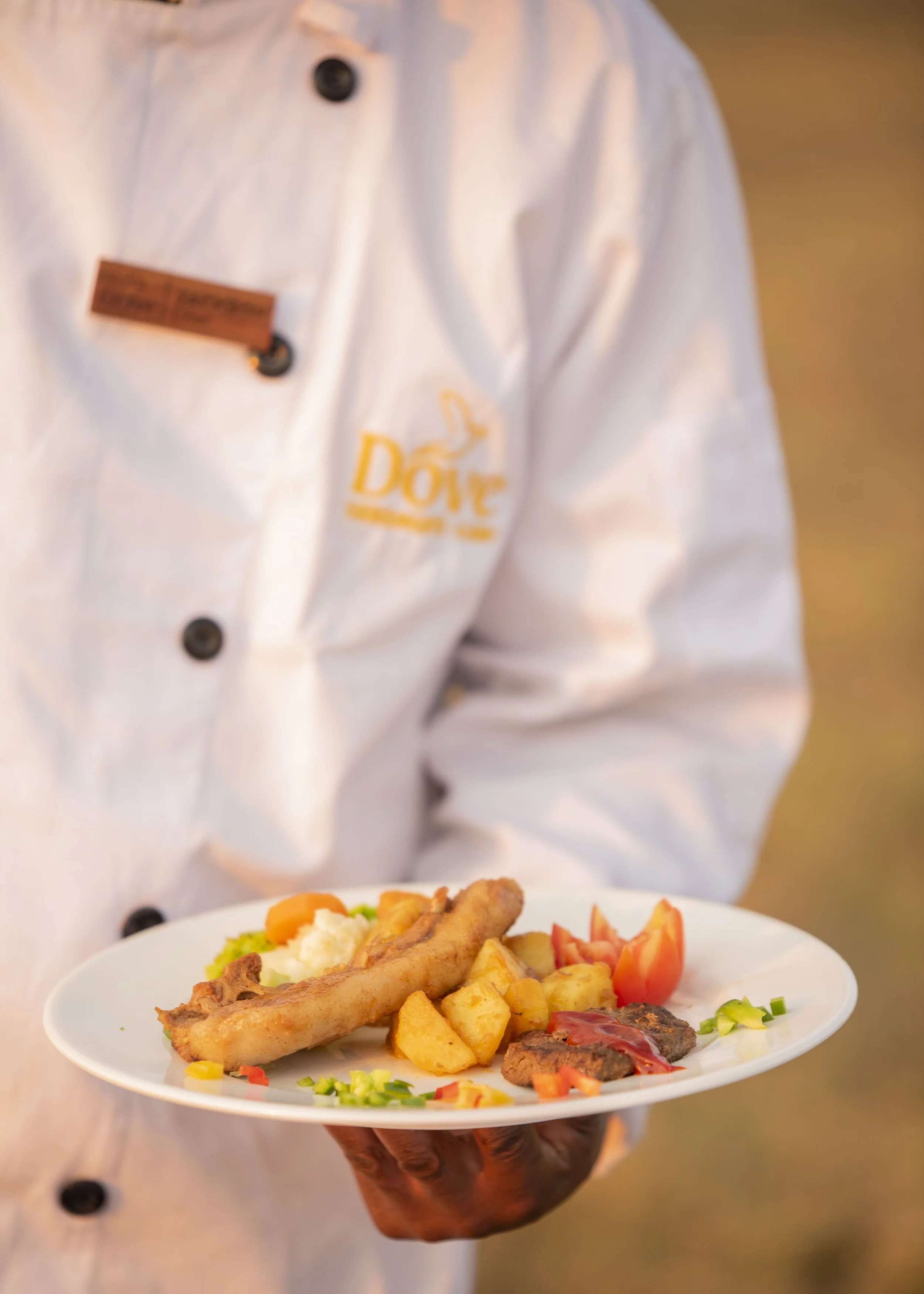 A chef in a white uniform holding a plate of food, including fried fish, potatoes, vegetables, and a small piece of meat with sauce, outdoors during sunset.