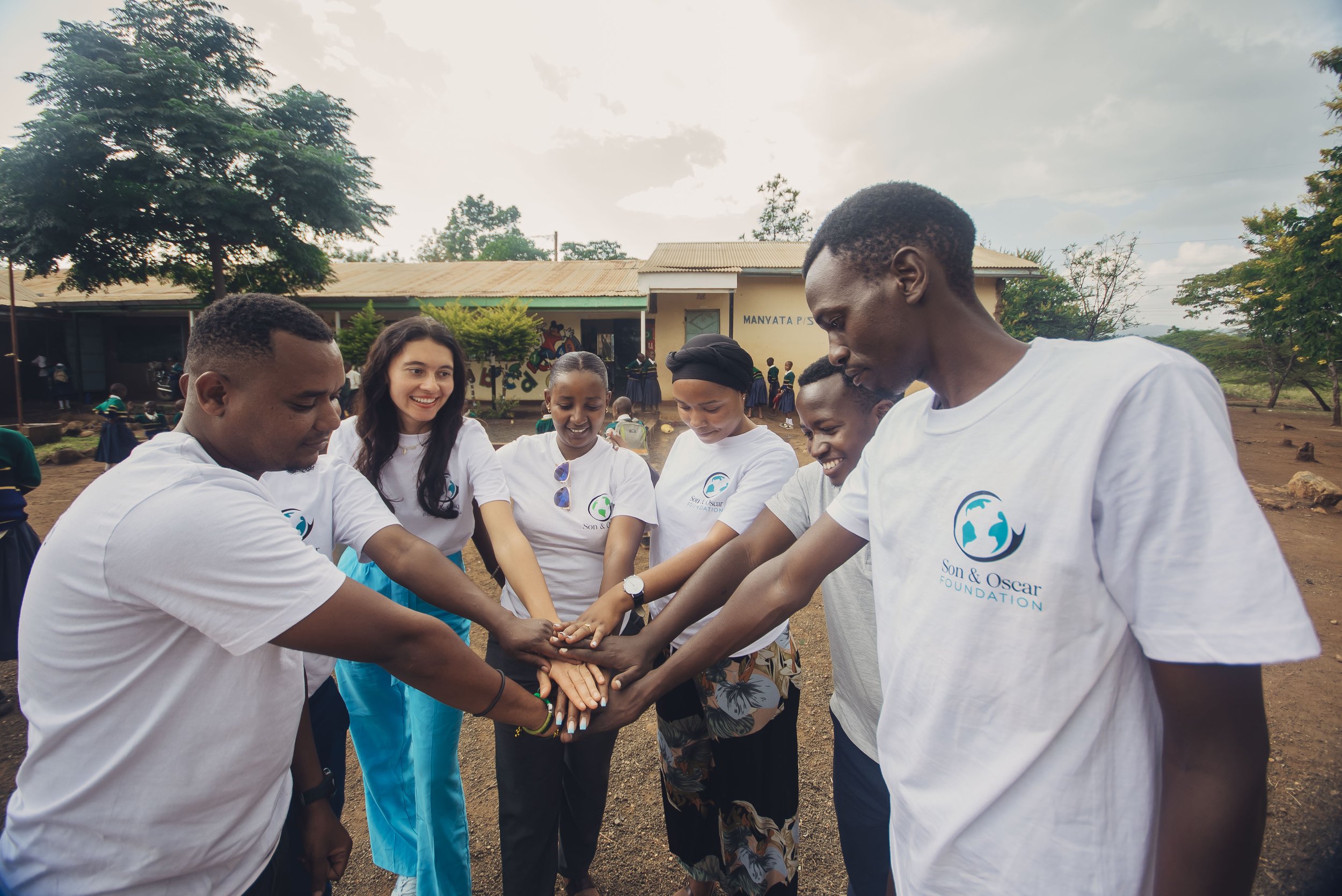 Group of diverse people in white t-shirts with the Son & Oscar Foundation logo, stacking hands together outdoors on a school playground, smiling and showing teamwork.