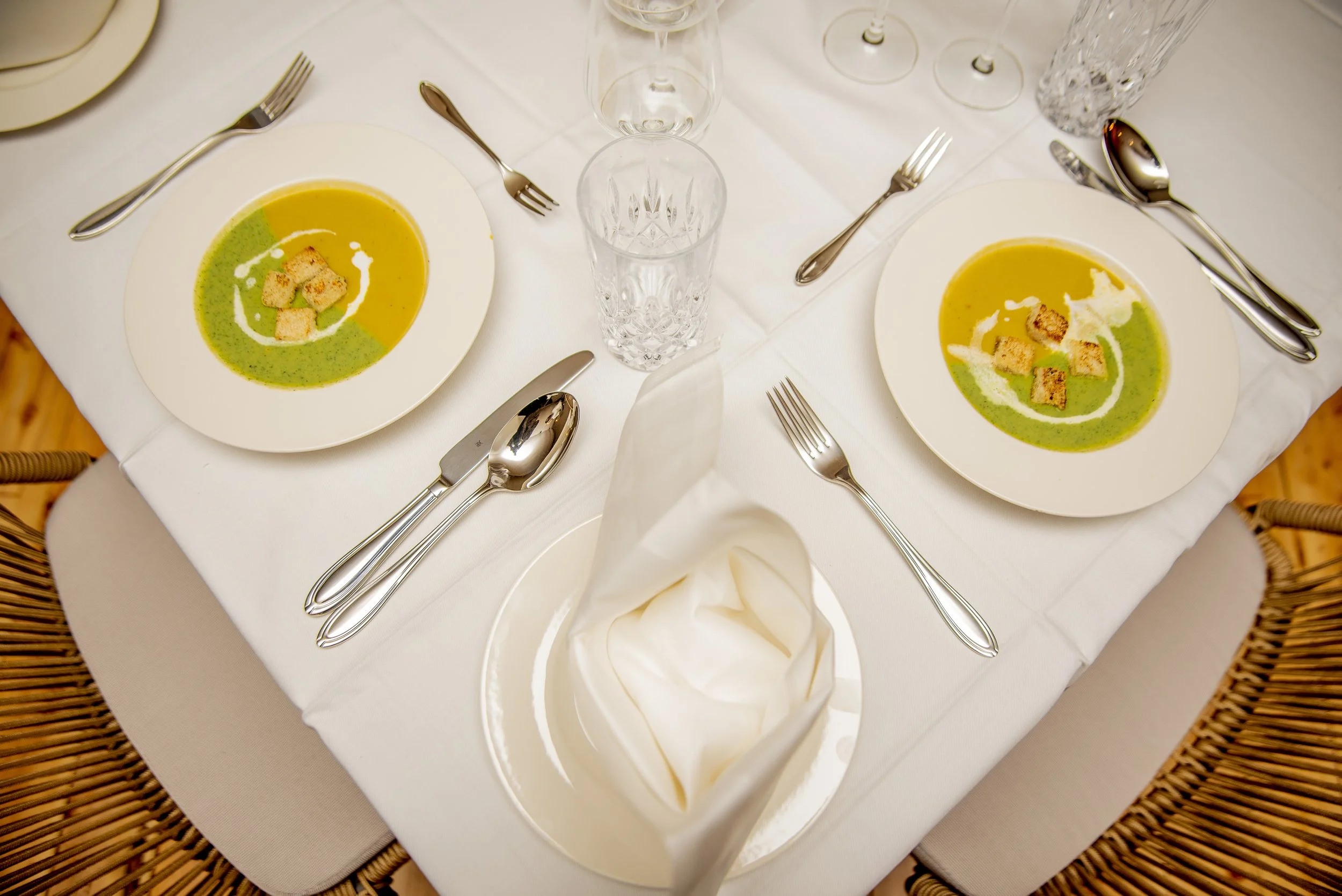 A formal dining table set for two, with white tablecloth, two bowls of green soup topped with croutons, two empty glasses, silverware, and a folded napkin in a bread plate.