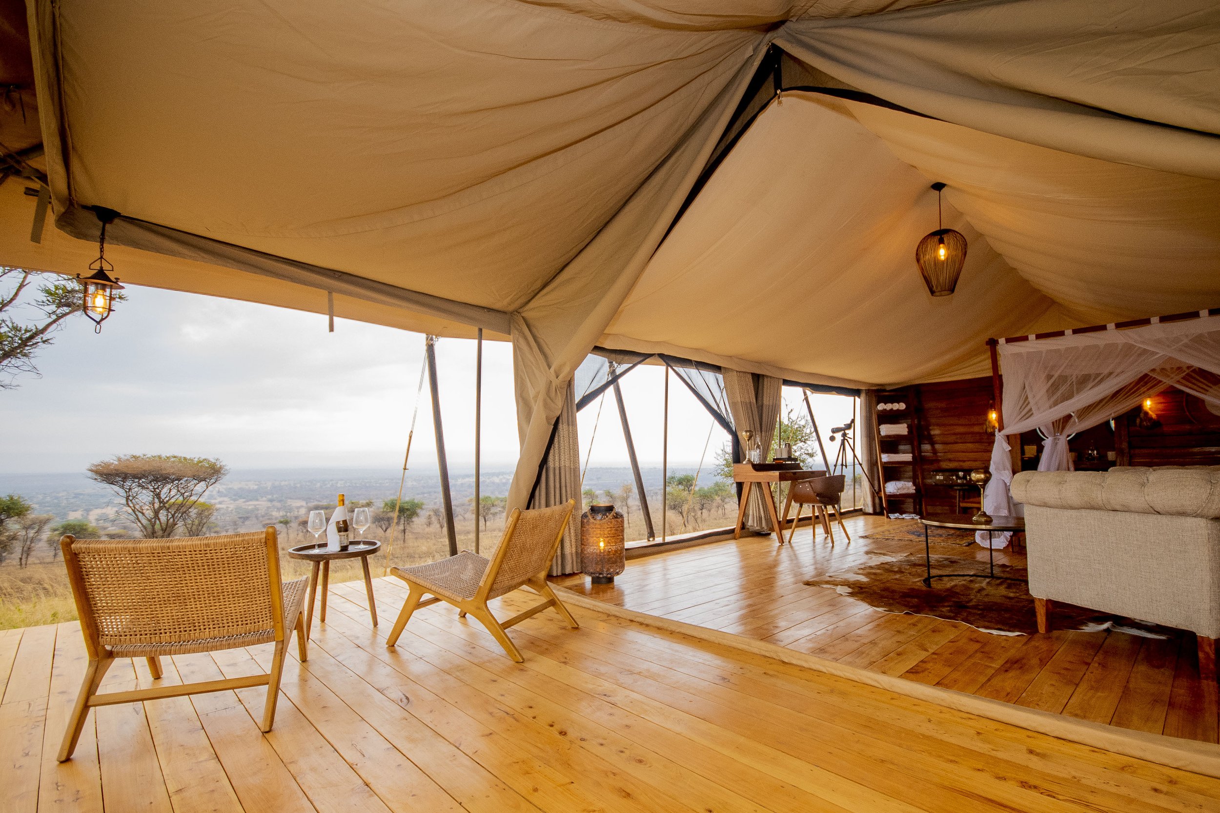 Interior of a luxurious glamping tent with a view of the savannah and trees outside, furnished with wooden chairs, a table with wine and glasses, a beige sofa, and warm lighting.