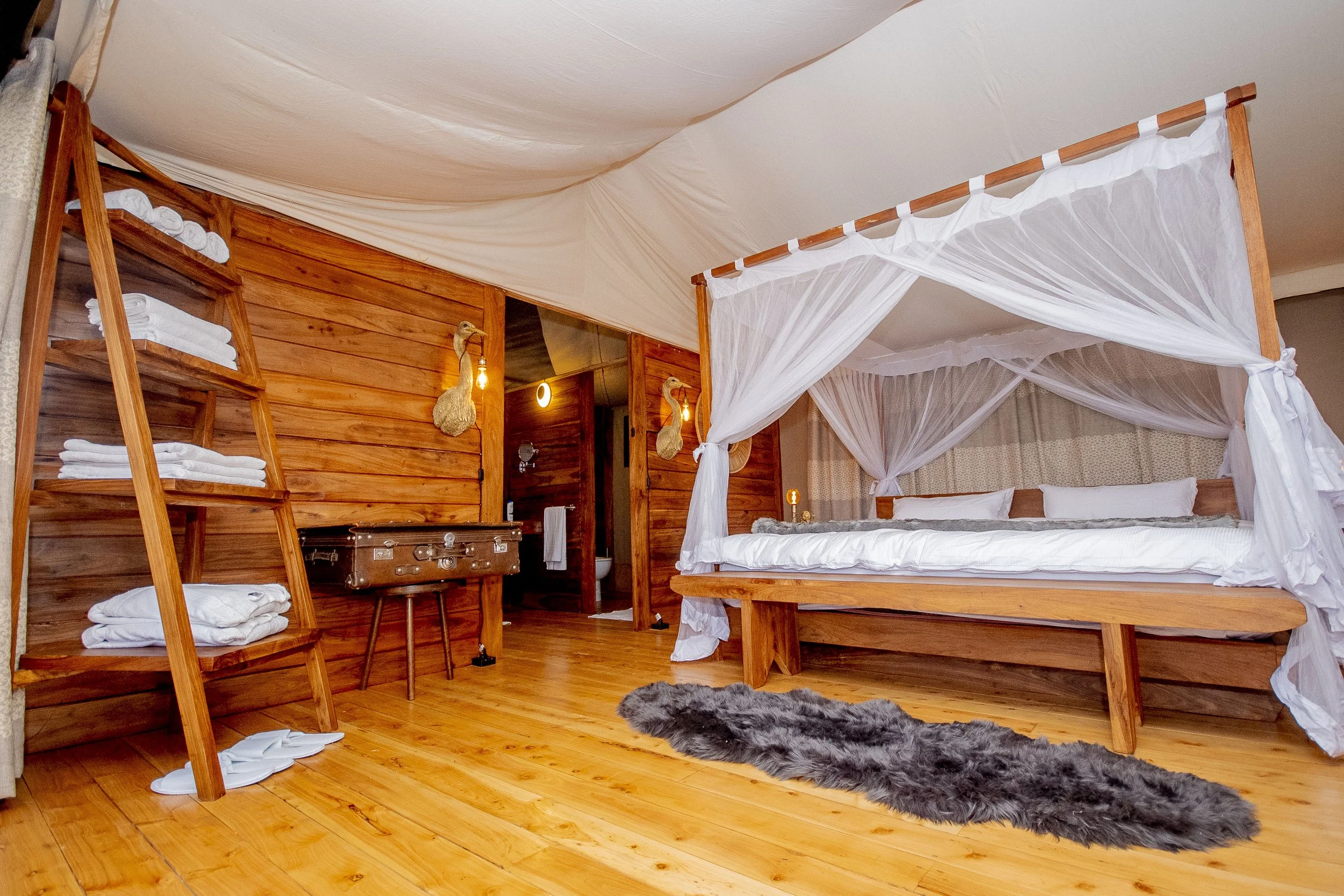 A cozy bedroom with a four-poster bed draped in white sheer curtains, a wooden ladder shelf with neatly folded towels, a furry gray rug on the wooden floor, and warm lighting accents on a wooden wall.