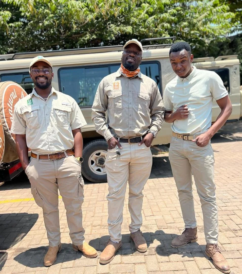 Three men standing outdoors on a paved area, posing for the photo. They are dressed in light-colored casual clothing and are smiling. There is a beige vehicle with large windows behind them, and green leafy trees in the background.