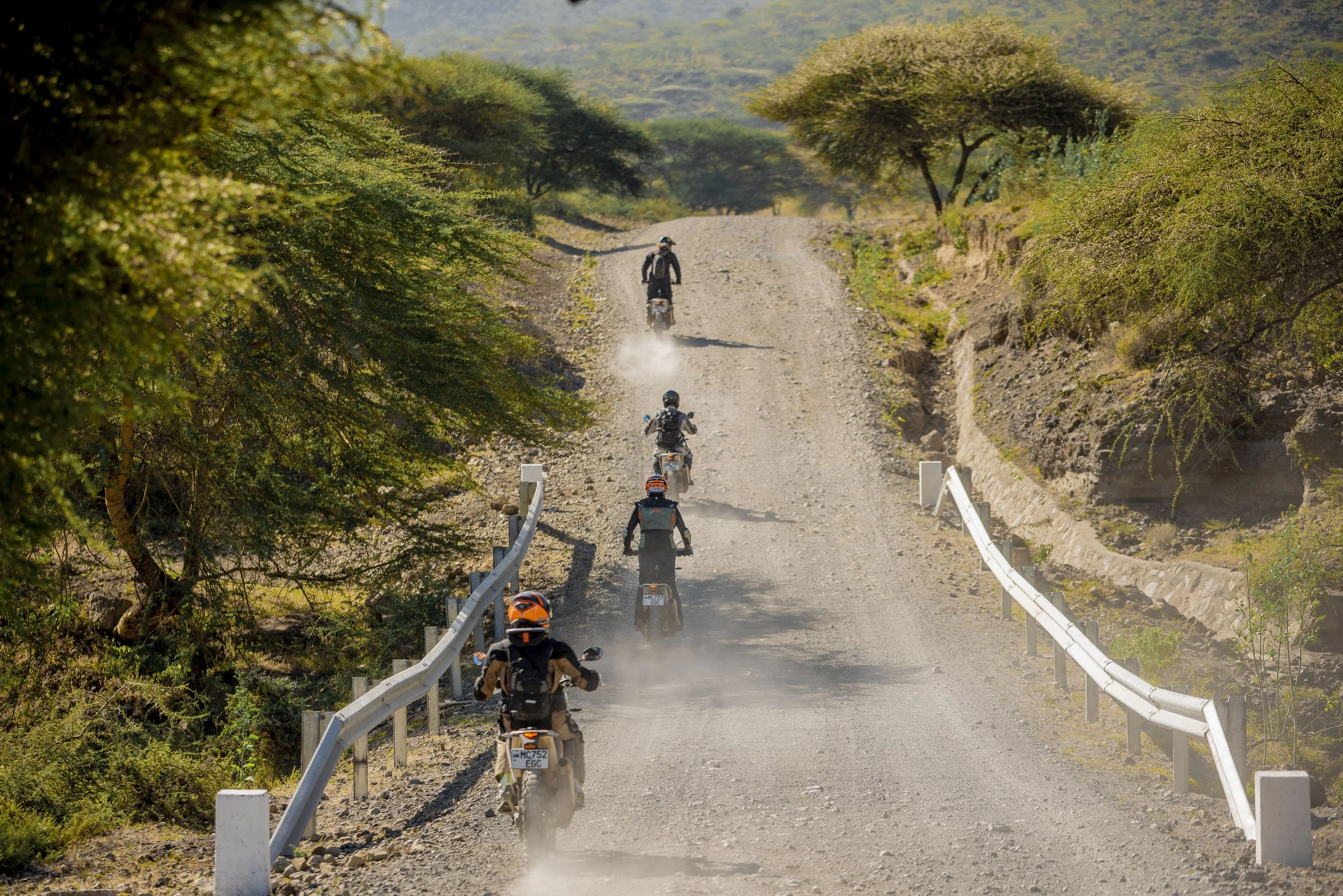 A group of five people riding dirt bikes on a dusty, gravelly off-road trail through a semi-arid landscape with sparse green trees and bushes, with hills in the background.