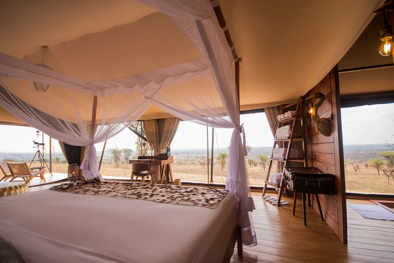 A bedroom with a large bed decorated with a leopard print blanket, mosquito netting, and large windows with a view of the savannah landscape, trees, and a cloudy sky.