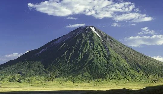 A tall, cone-shaped volcano with green slopes and a snow-capped crater, under a partly cloudy sky.