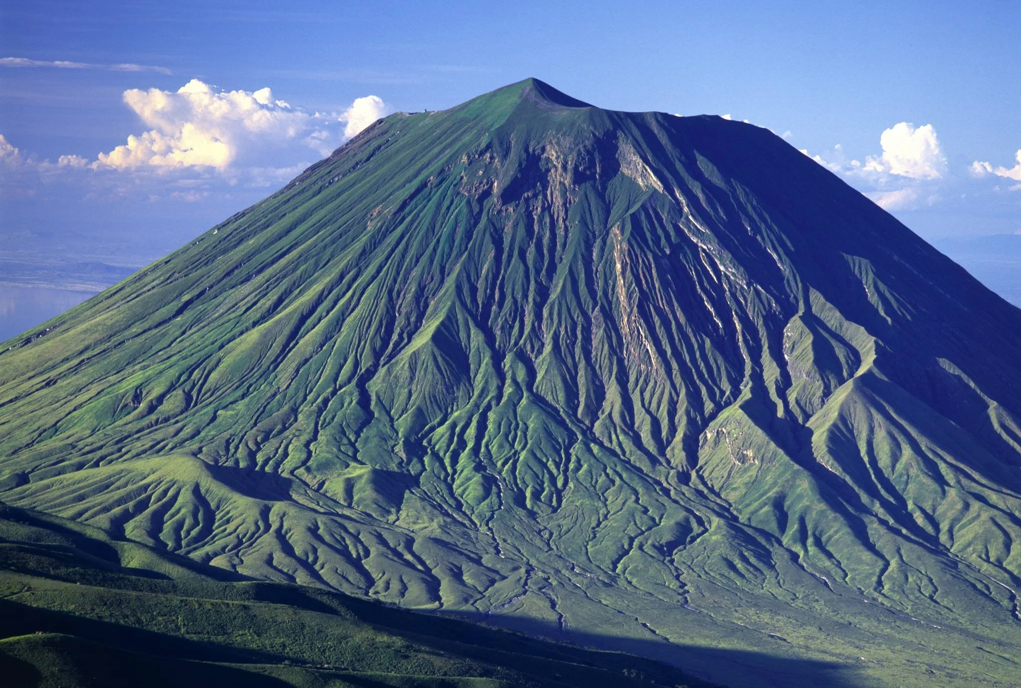 A snow-capped mountain with lush green slopes and deep ridges, against a partly cloudy sky.