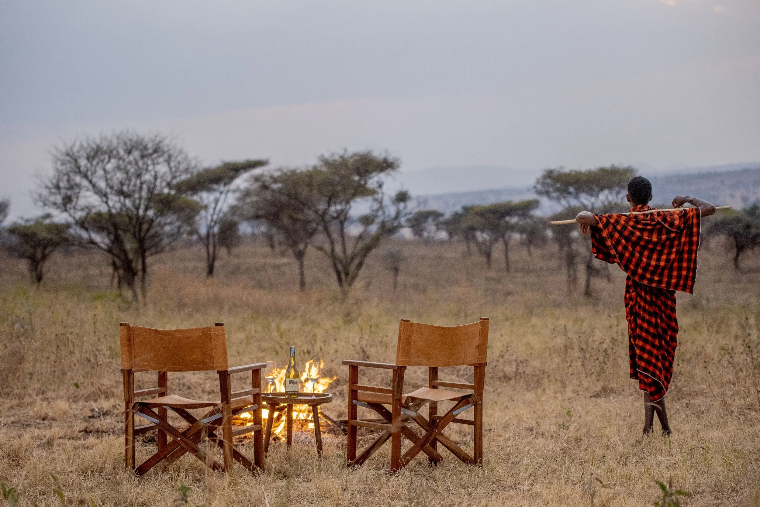 A person dressed in traditional Maasai clothing walks across the savannah holding a spear, with two chairs and a small table with drinks and a fire in the foreground, and scattered trees in the background.