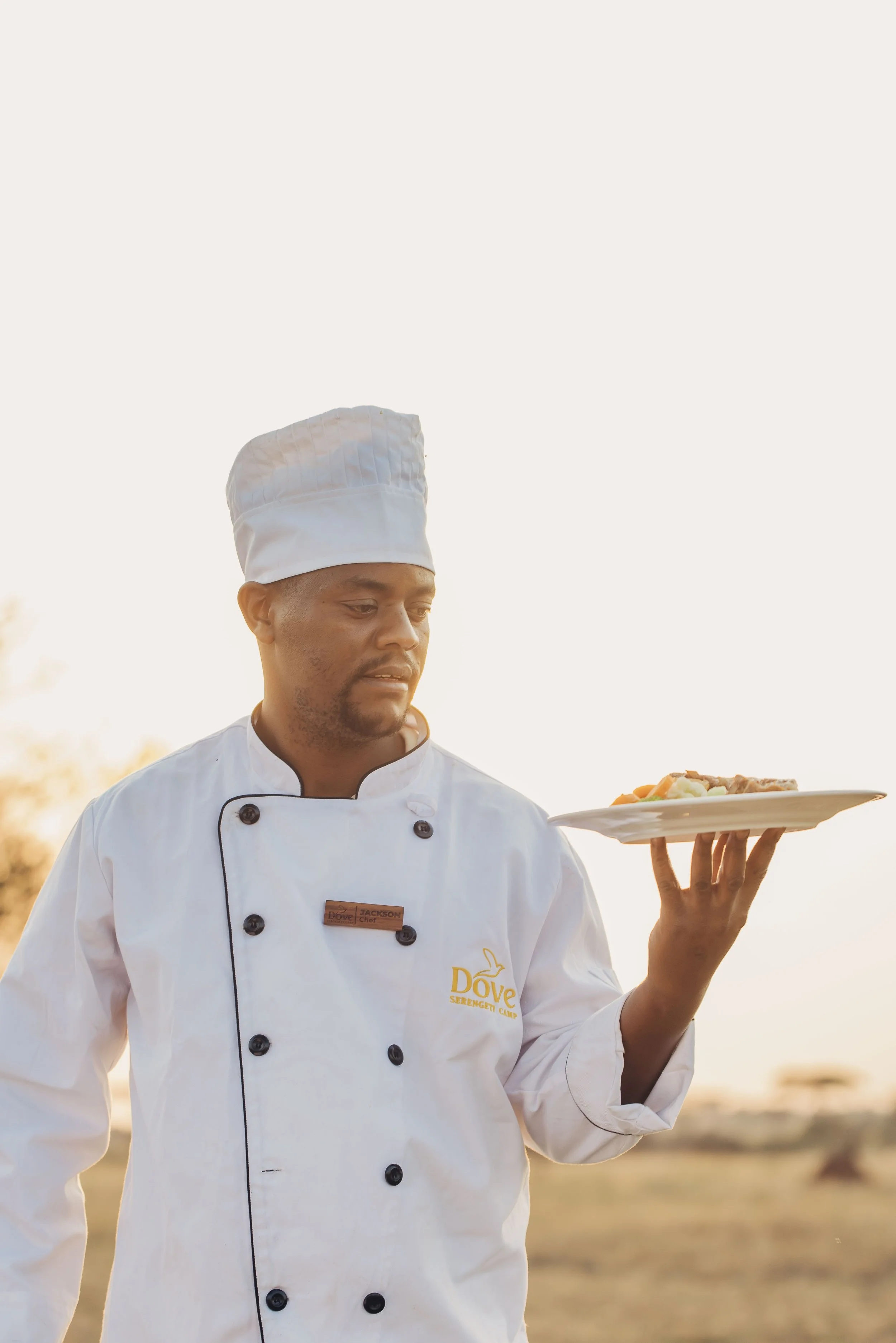 A chef in a white uniform and tall chef hat holding a plate of food outdoors during sunset.