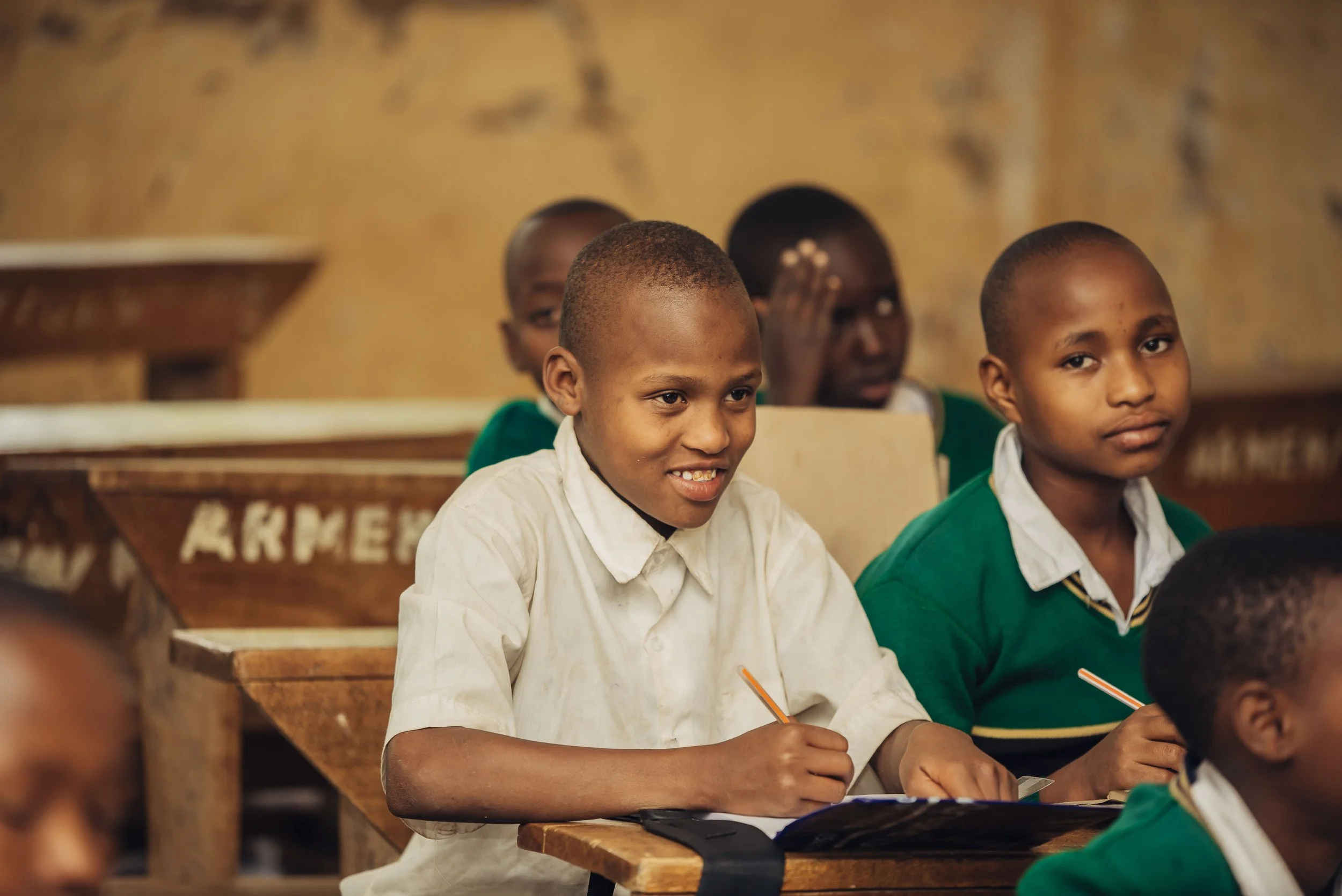 Young students sitting at desks in a classroom, with some smiling and others serious, some holding pens.