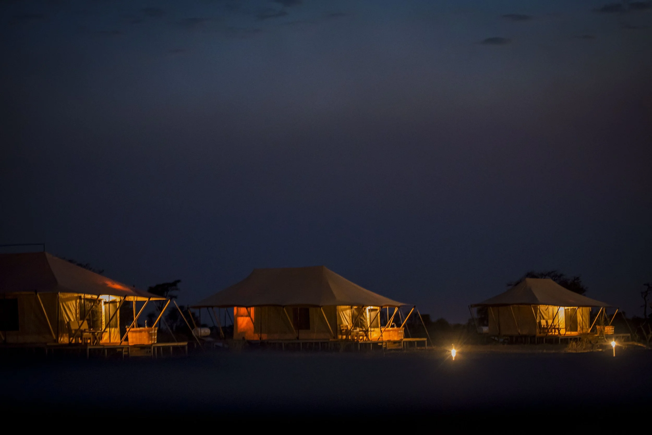 Three illuminated tents on a dark night, with a crescent moon and starry sky overhead, surrounded by outdoor lighting.