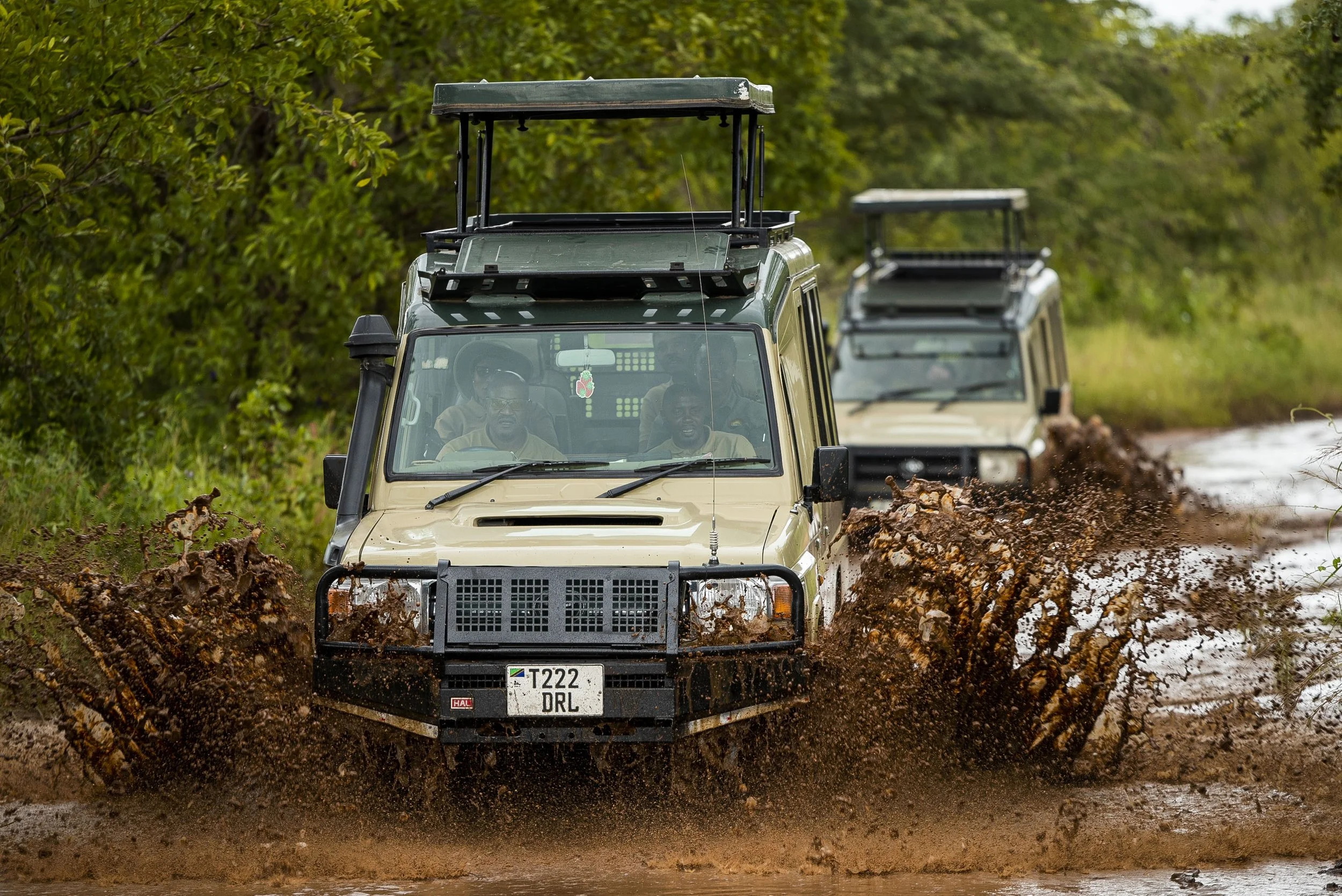 Two off-road vehicles driving through a muddy trail with splash and greenery on the sides.