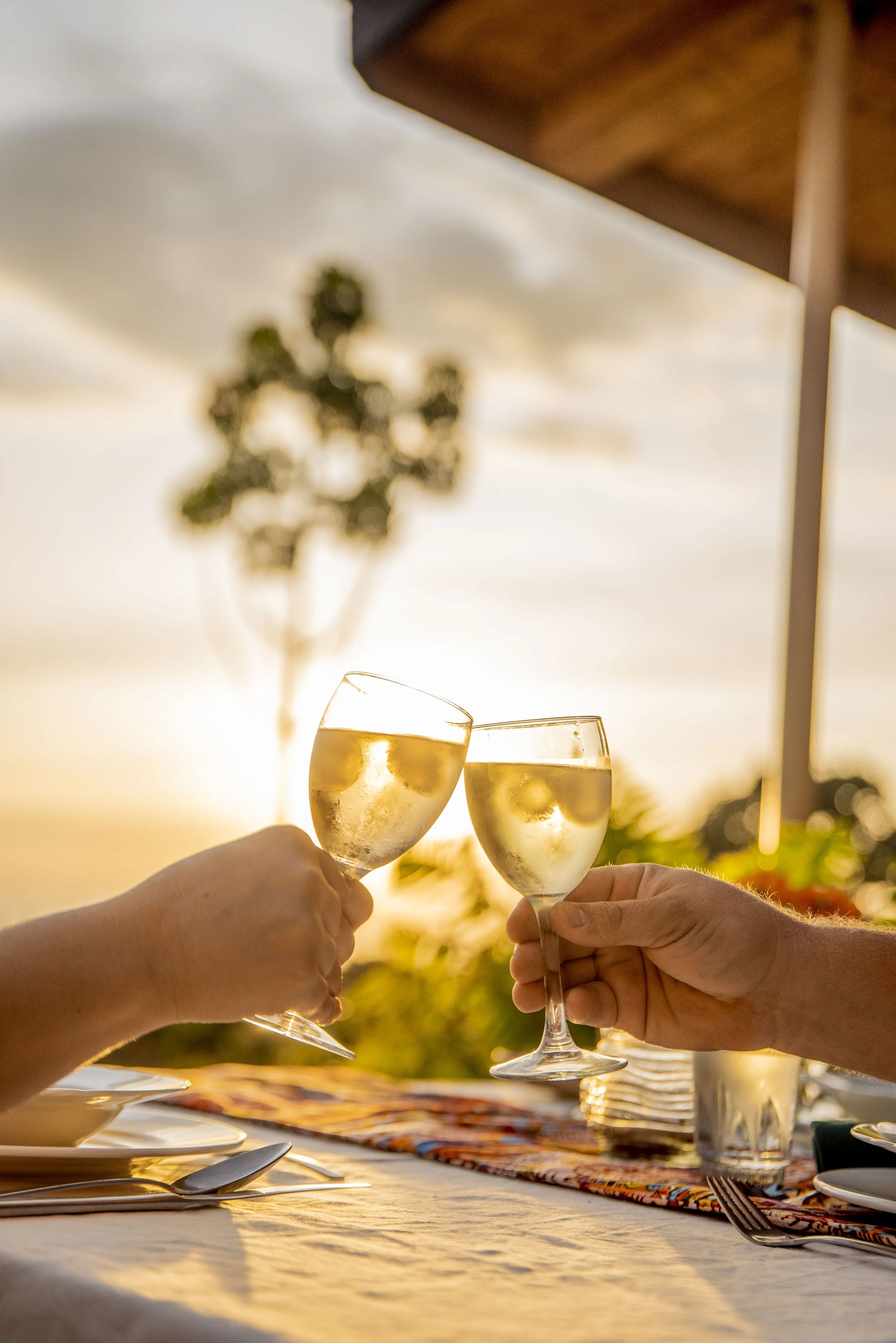 Two people clinking glasses of white wine or champagne during sunset on an outdoor dining table.
