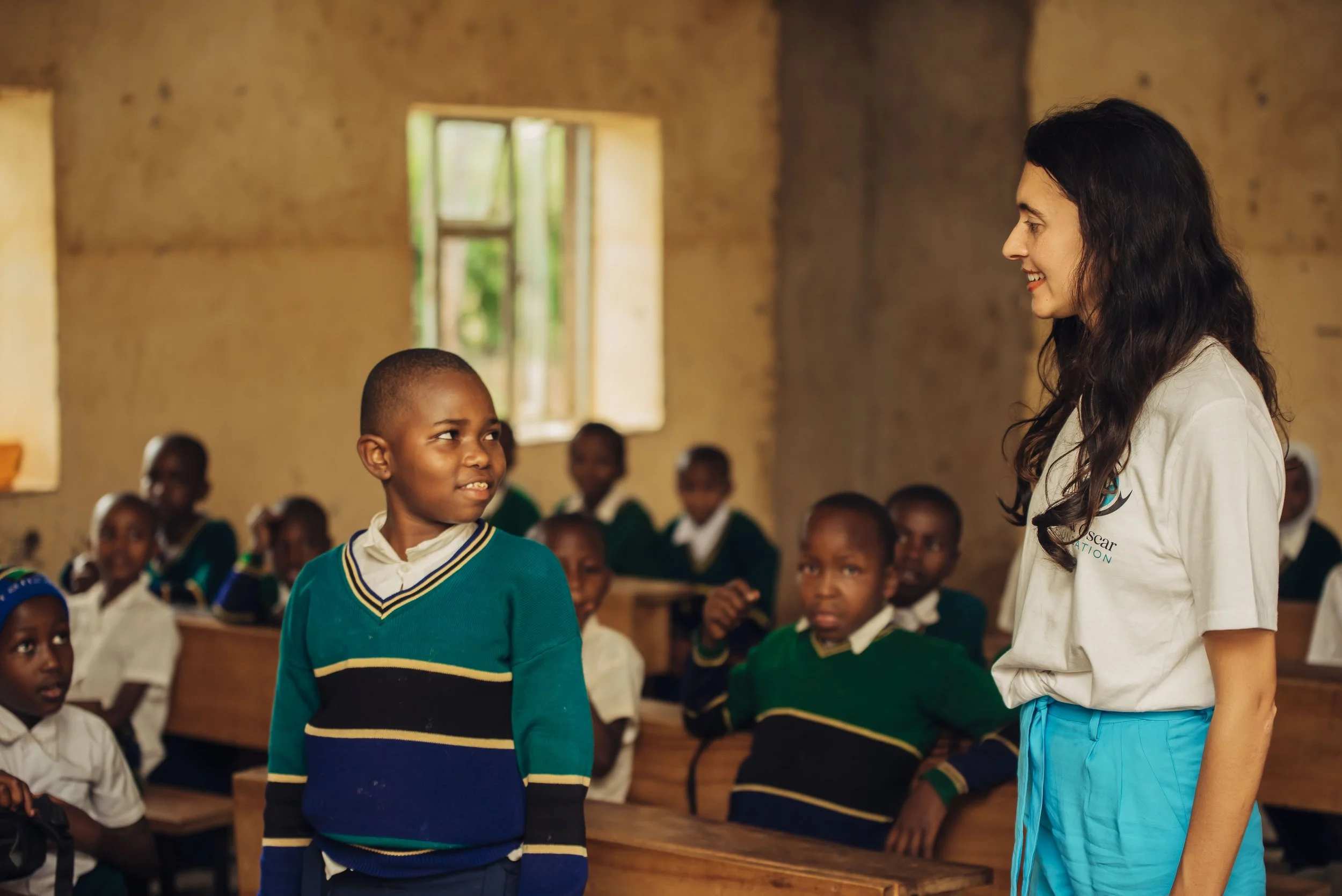 A young boy in a school uniform standing and talking to a woman in a classroom of children, some sitting at desks, with a wall and window in the background.