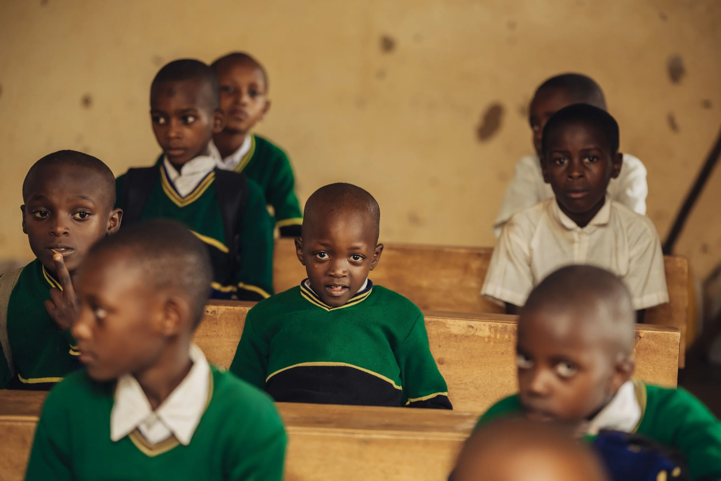 Young students sitting in a classroom, wearing green and white school uniforms, some students are looking at the camera while others are looking elsewhere.