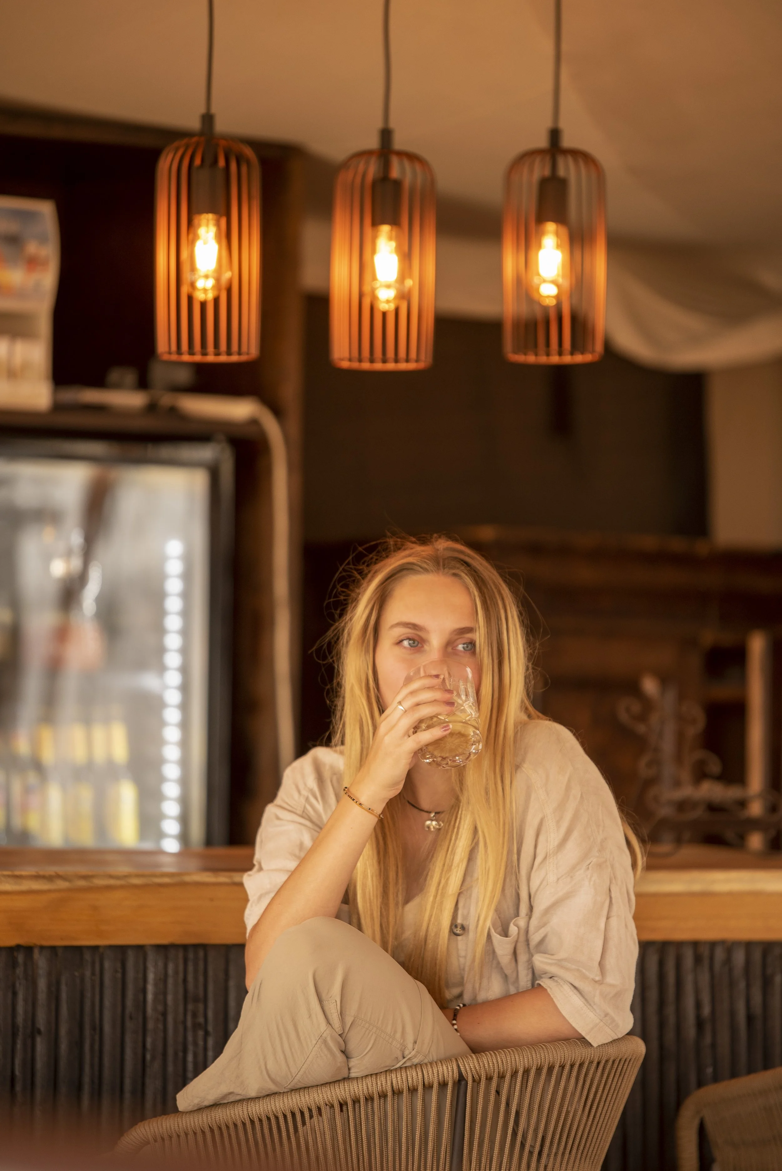 A young woman with long blonde hair sitting in a cozy cafe or restaurant, drinking from a glass, with three hanging pendant lights above her.