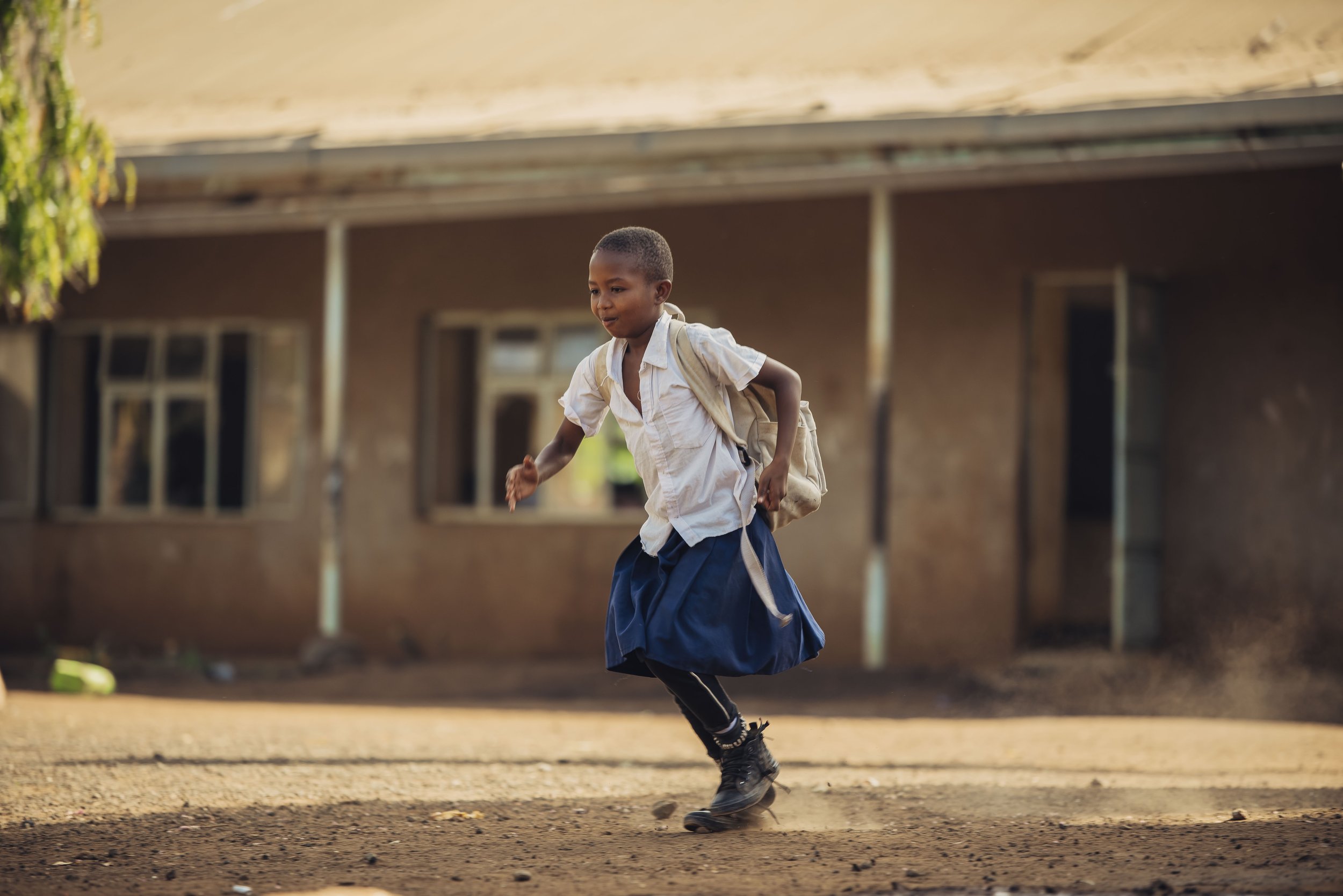 A young boy in school uniform running on a dirt ground in front of a building with open windows.