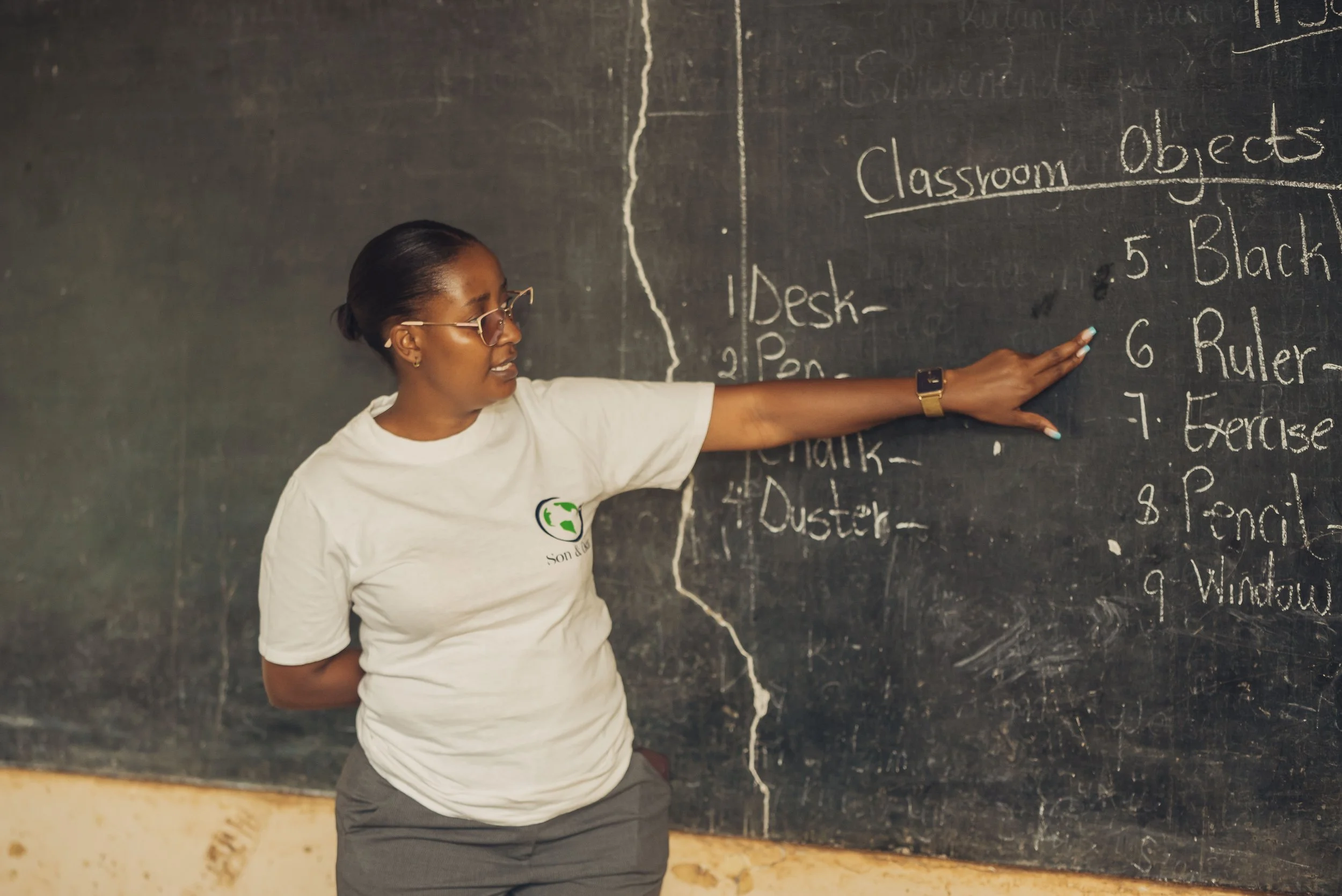A woman pointing at a chalkboard with a list of classroom objects written on it.