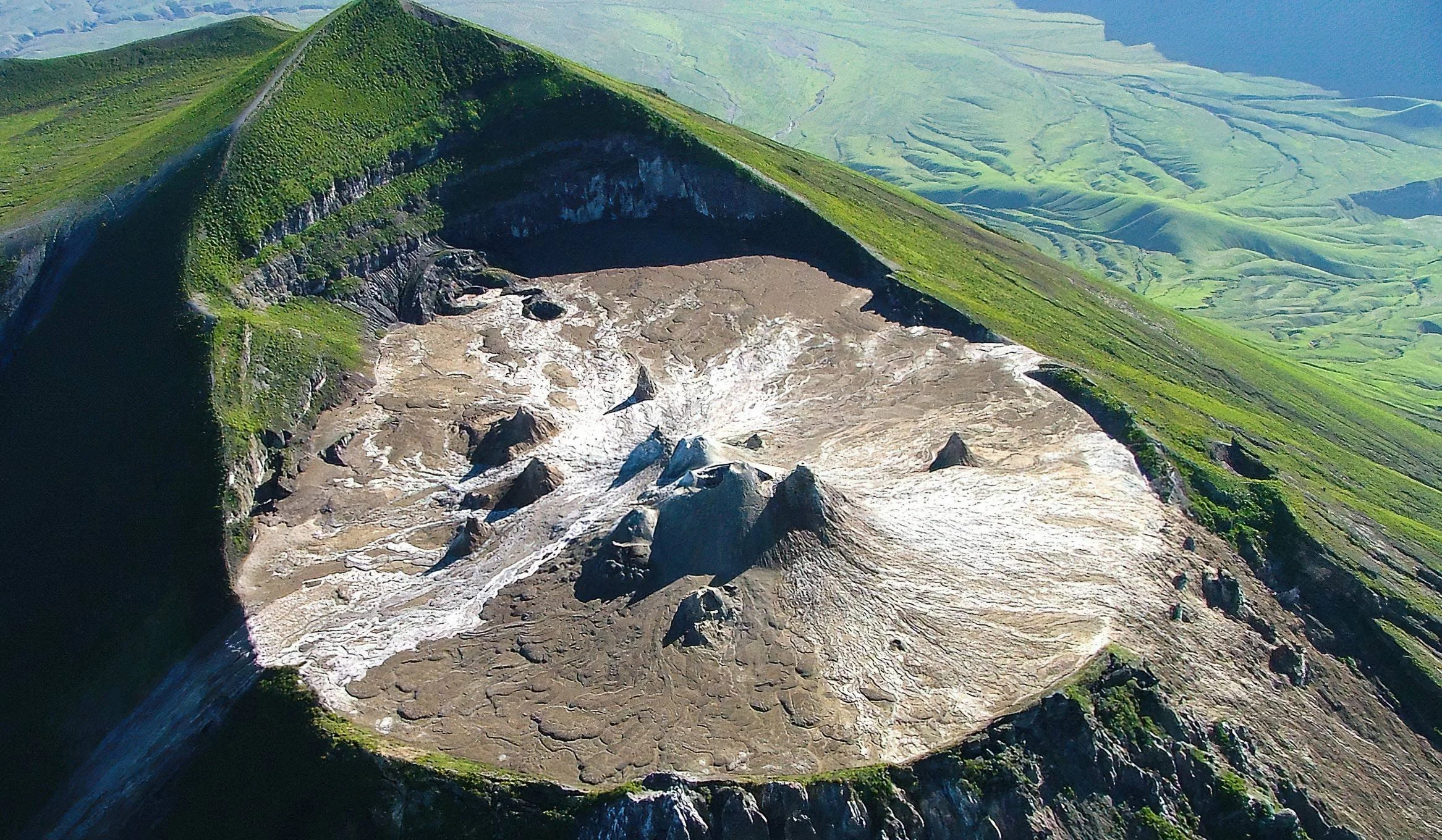 A volcanic crater with a glacier or snow cover inside, surrounded by green mountains and landscape.