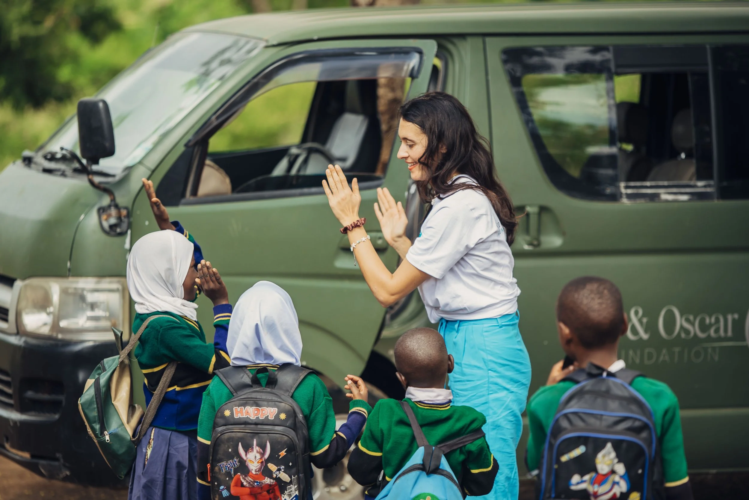 A woman in a white shirt and blue pants interacts with four children in green school uniforms, standing in front of a green vehicle with a logo that reads 'Oscar Foundation', on a grassy outdoor area.