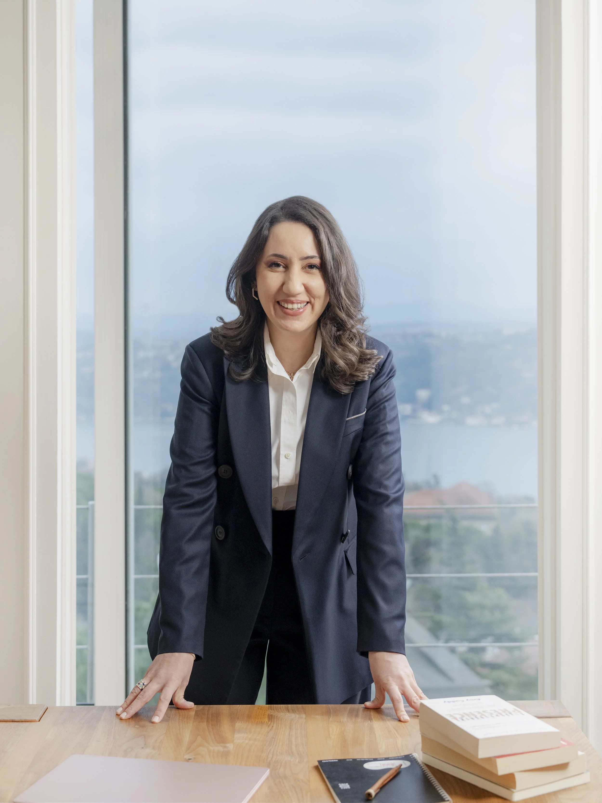 Geniş pencereleri ve su ile dağların manzarasına sahip aydınlık bir odada, ahşap bir masaya eğilmiş, lacivert takım elbiseli bir kadın kameraya gülümsüyor.