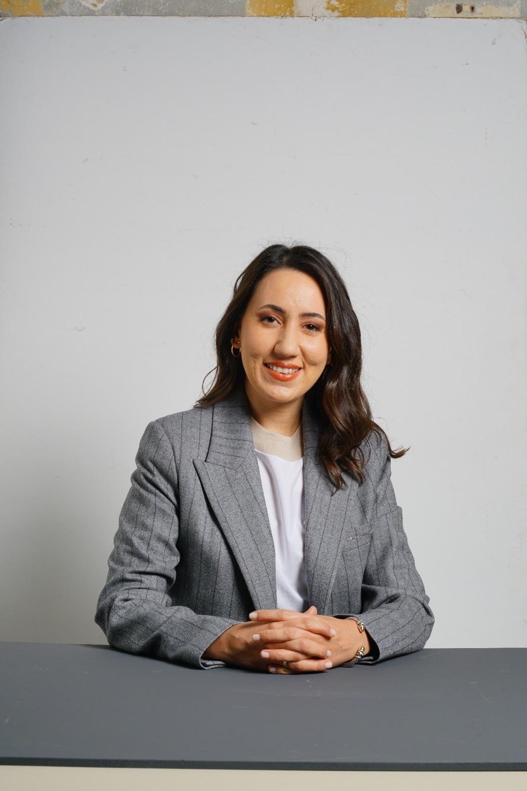 Omuz uzunluğunda dalgalı kahverengi saçlı, beyaz bluz üzerine gri çizgili blazer giymiş, gülümseyen ve düz beyaz bir duvarın önünde gri bir masada oturan bir kadın.