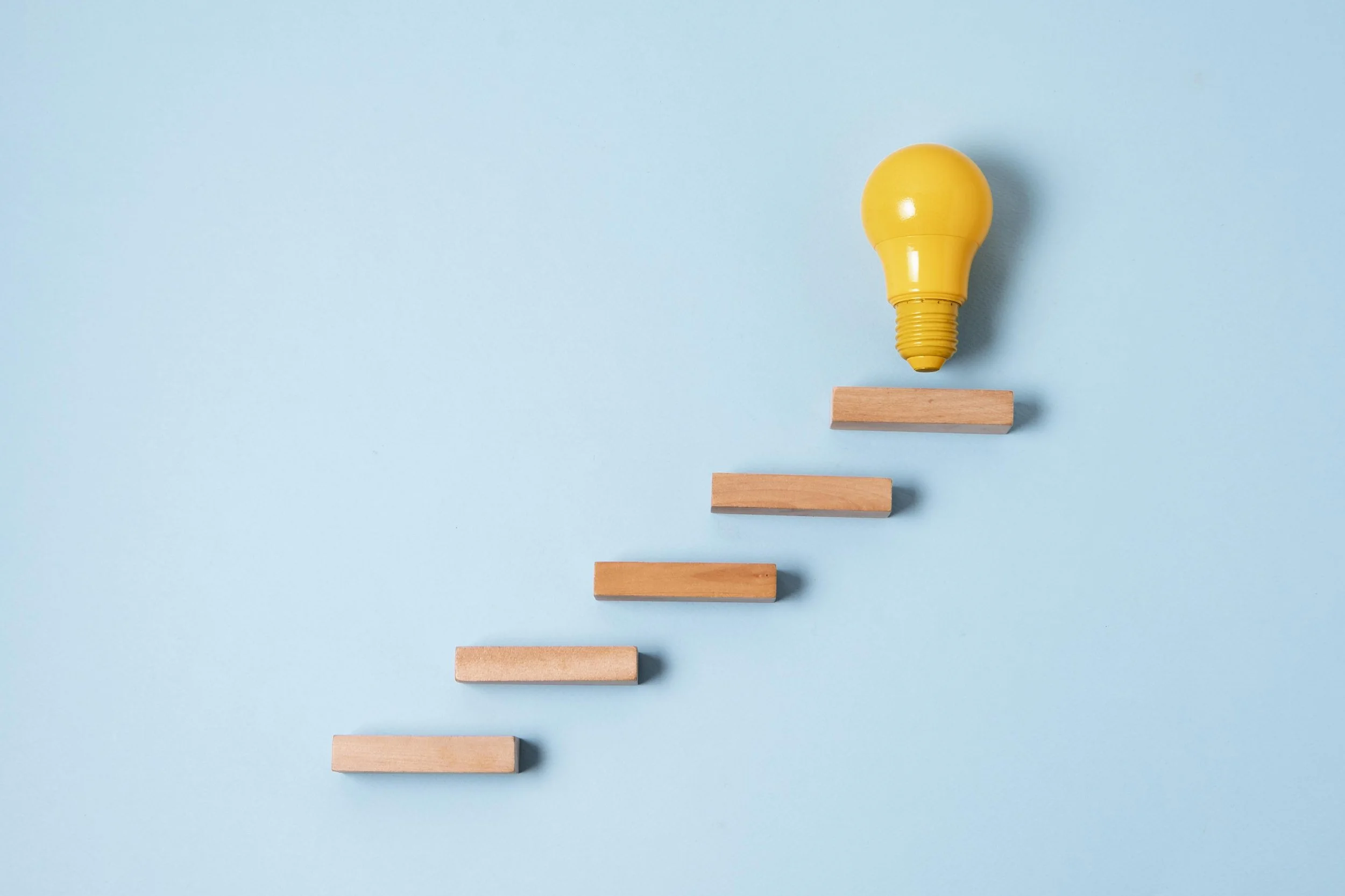 Açık mavi bir arka plan üzerinde üç tahta bloğun üzerine yerleştirilmiş sarı bir ampul, fikirleri ve ilerlemeyi simgeliyor.
