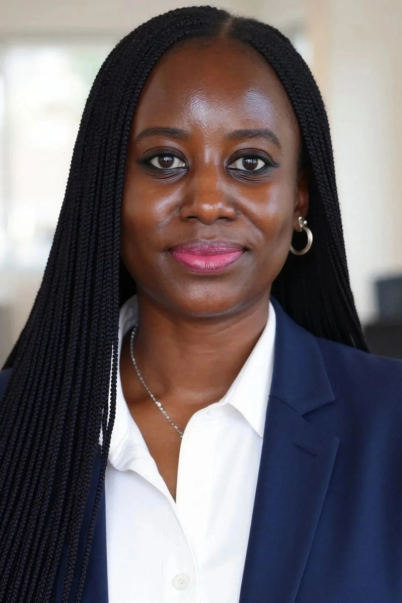 Close-up of an African American woman with braided hair, wearing a dark blazer over a white shirt, silver hoop earrings, and a silver necklace, smiling softly.