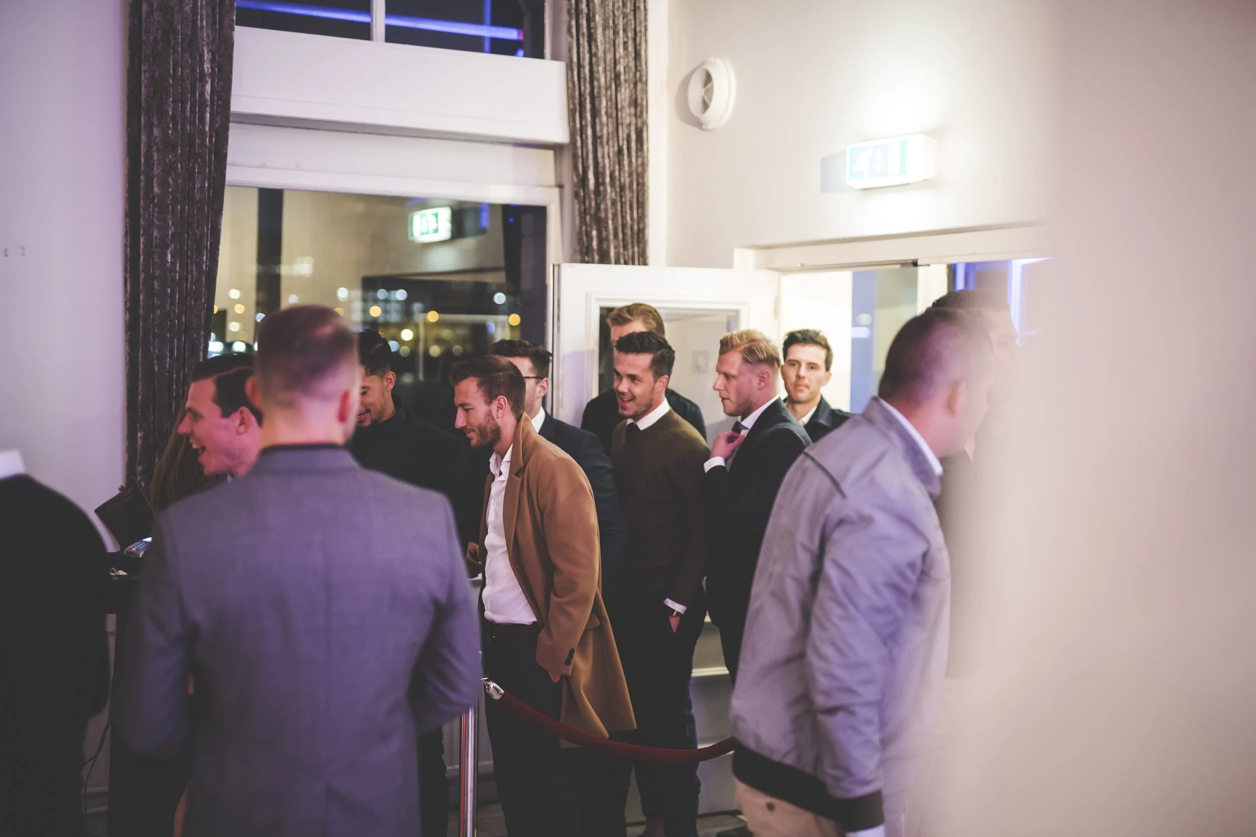 Groep mannen wacht in rijen bij de ingang van een evenement in een binnenruimte met grote ramen en gordijnen, binnen uitzicht op stadslichten 's nachts.