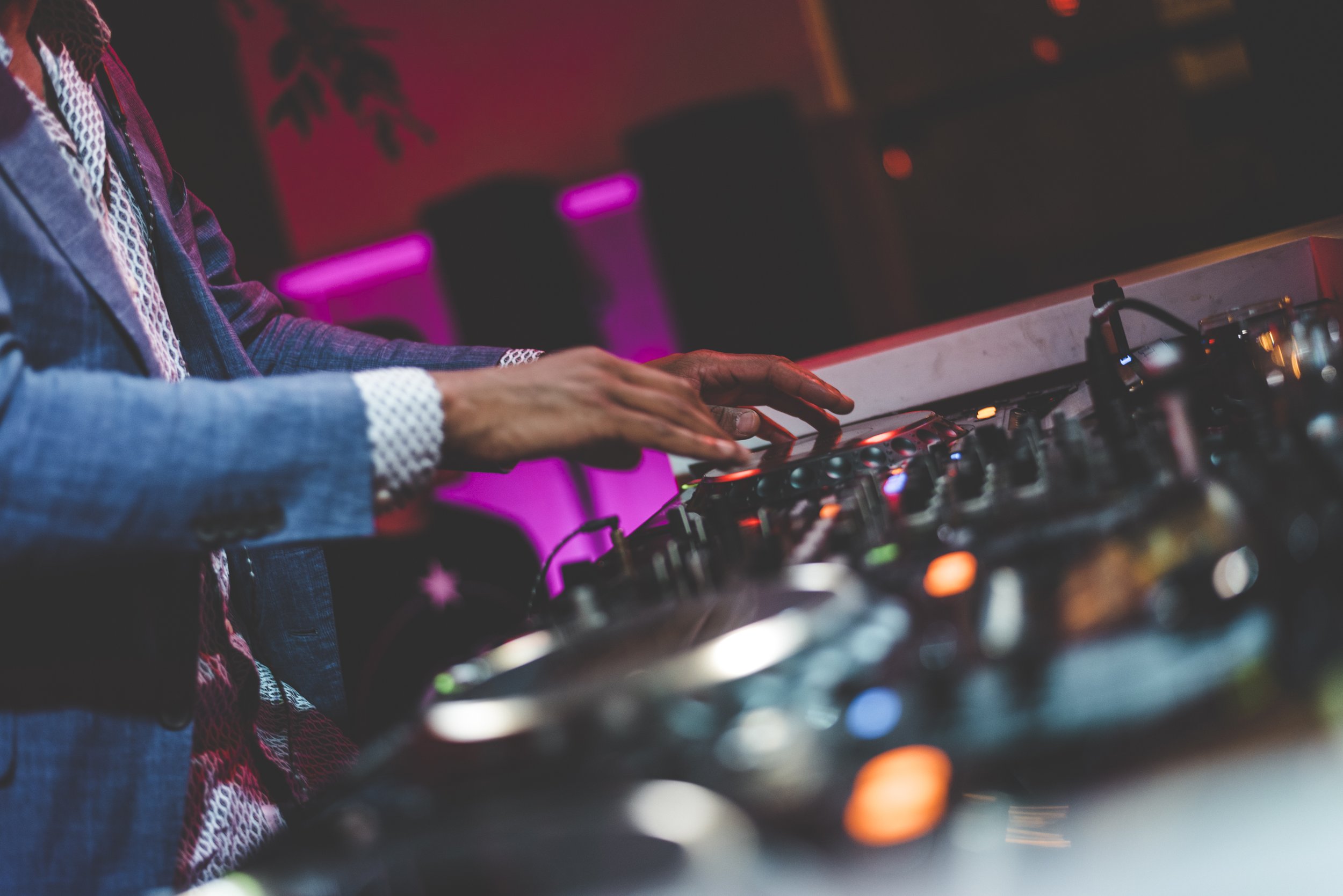 DJs handen bedienen mengpaneel met gekleurde knoppen en schuifregelaars in een club met roze en blauwe verlichting.