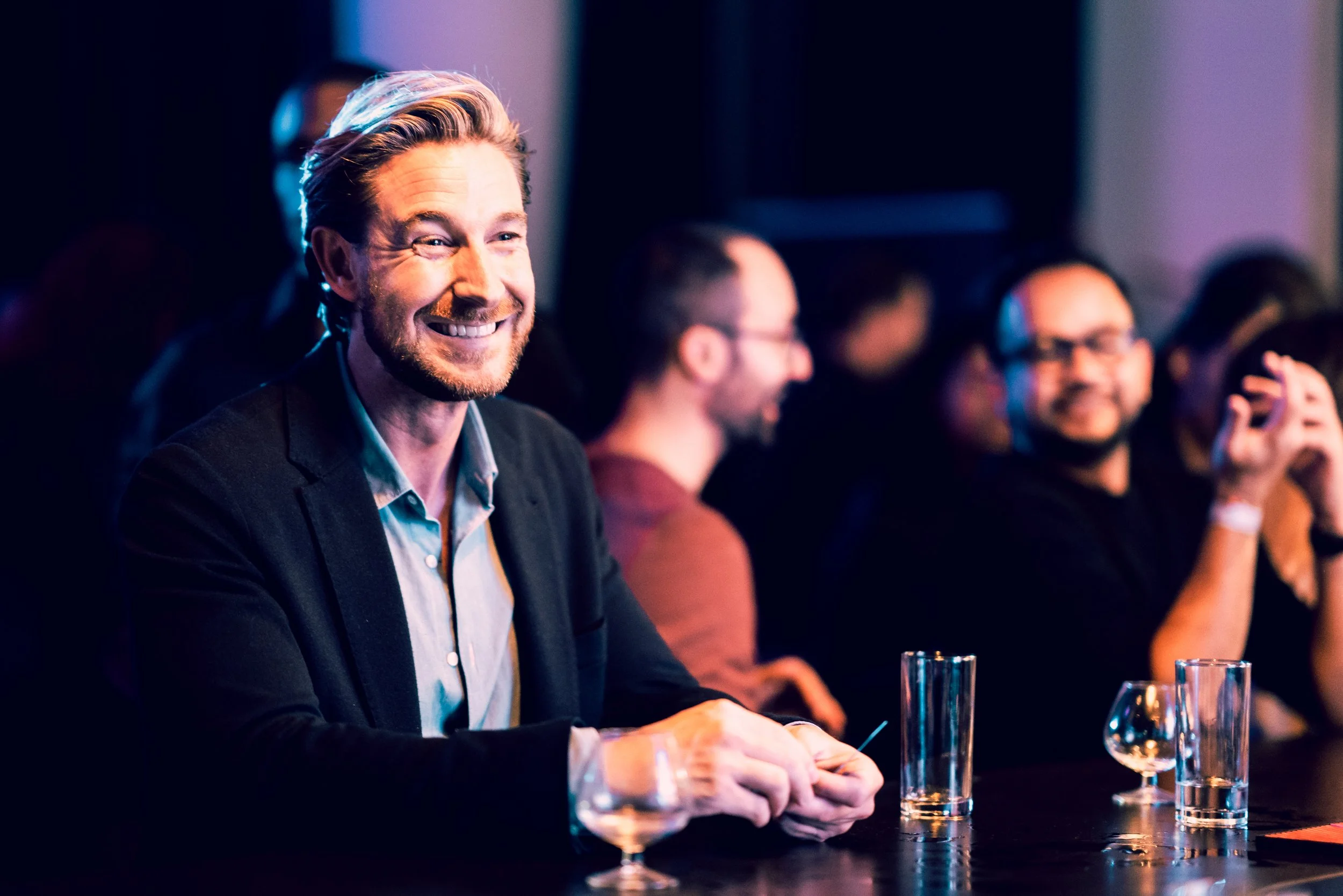 Vrolijke man met blond haar en een lichte baard lacht op een feestje of congres, omringd door andere mensen die ook genieten.
