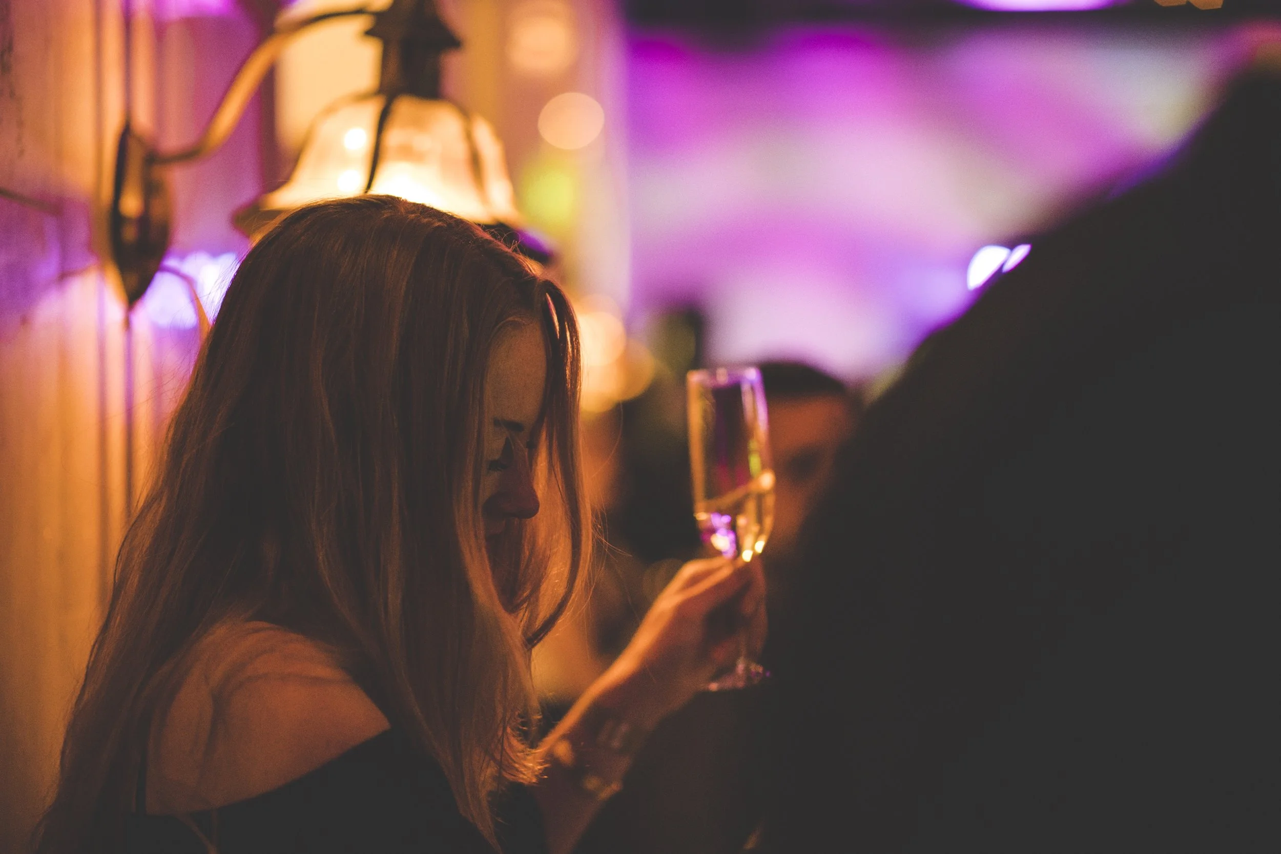 Een vrouw met lang haar houdt een glas champagne vast in een donkere, kleurrijke kamer.